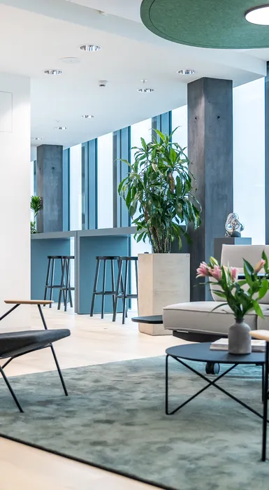 Gemütliche Sitzecke mit Sesseln und Sofa, flankiert von großen Pflanzen und Blick auf hohen Barbereich am Fenster.