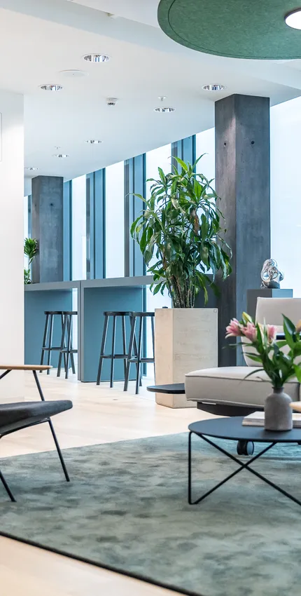 Gemütliche Sitzecke mit Sesseln und Sofa, flankiert von großen Pflanzen und Blick auf hohen Barbereich am Fenster.