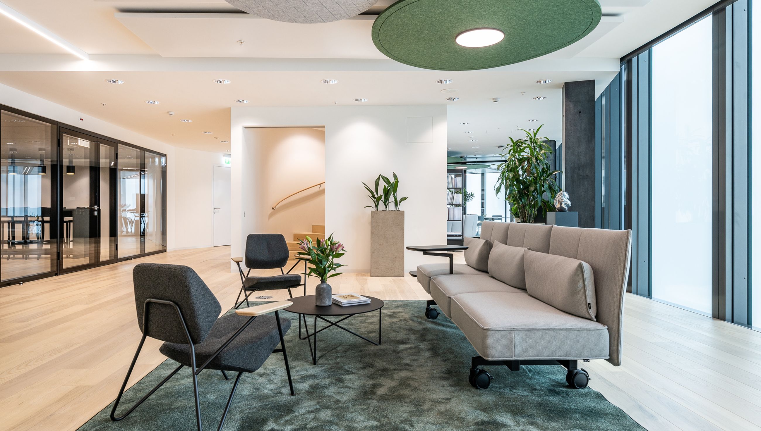 Offene Loungezone mit Sofas und Sesseln auf Teppich, angrenzend an verglaste Meetingräume und helle Arbeitsbereiche.