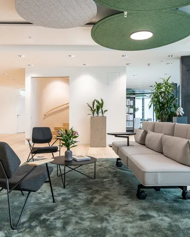 Offene Loungezone mit Sofas und Sesseln auf Teppich, angrenzend an verglaste Meetingräume und helle Arbeitsbereiche.