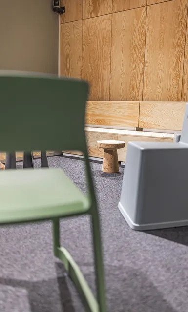 Grüner Stuhl im Vordergrund, Sitzbank vor Holzwand im Hintergrund.