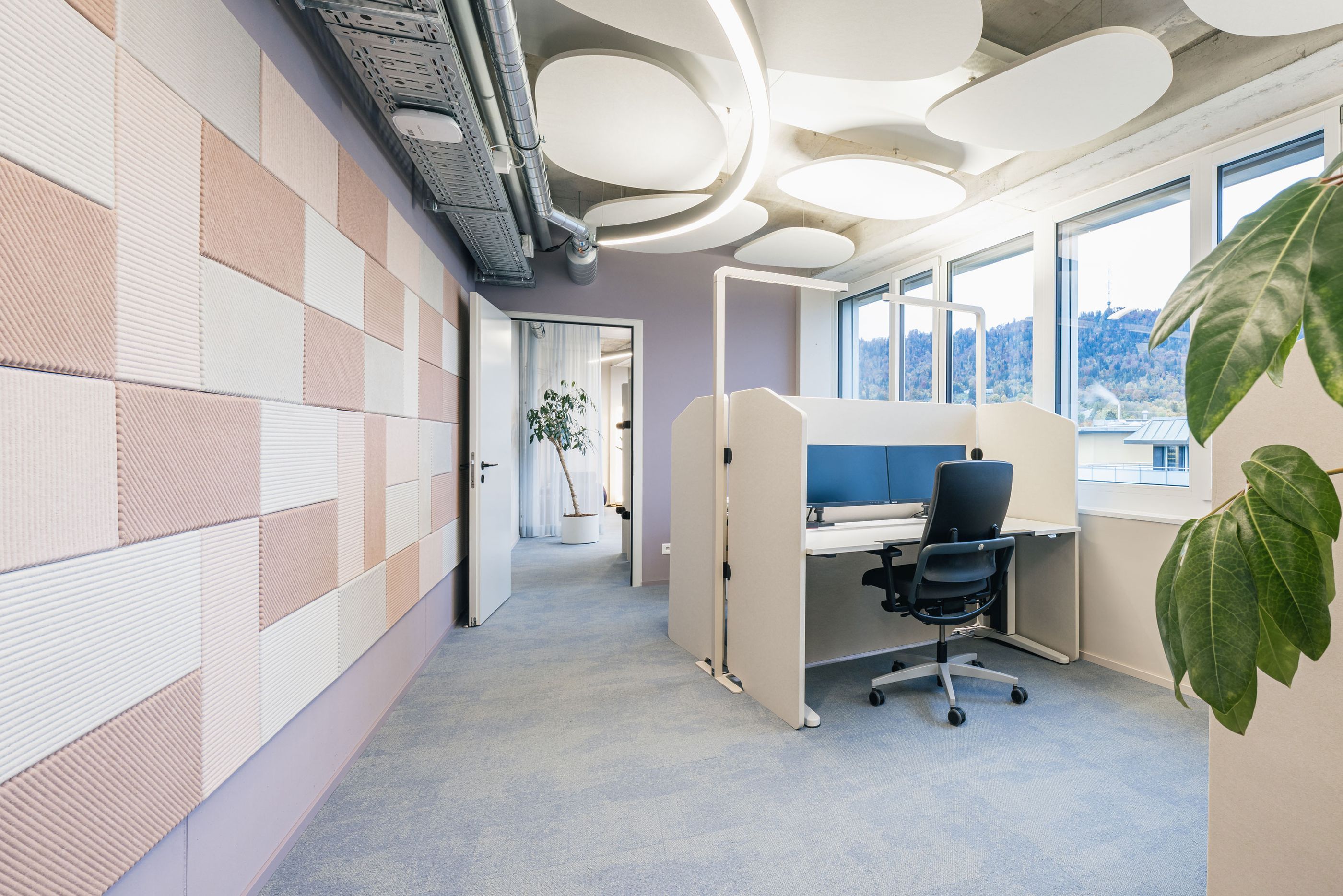 Heller Bürobereich mit pastellfarbenen Akustikpaneelen, Einzelarbeitsplatz und großen Fenstern.