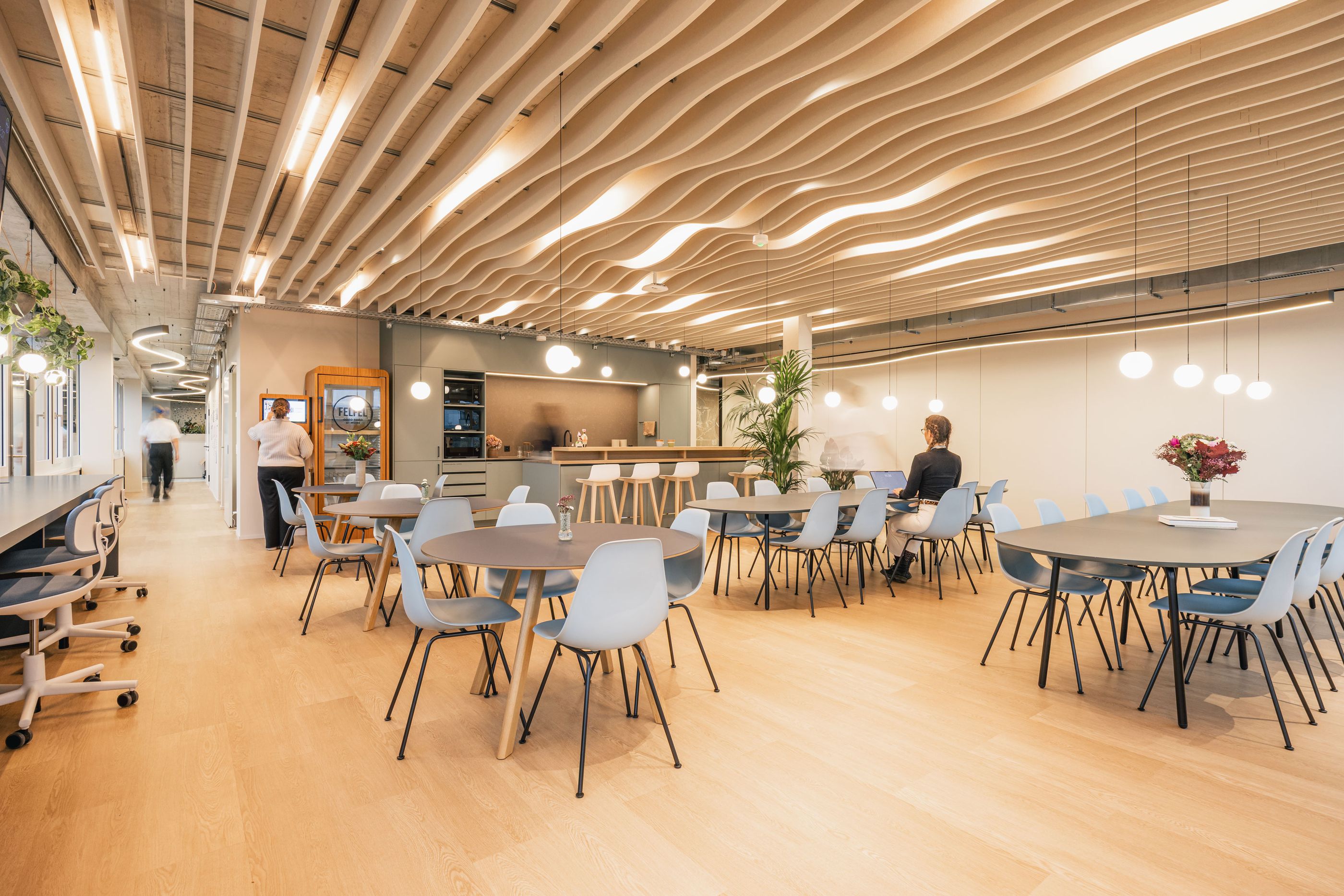 Großer Aufenthaltsbereich mit Tischen, hellblauen Stühlen, offener Küche und wellenförmiger Decke.