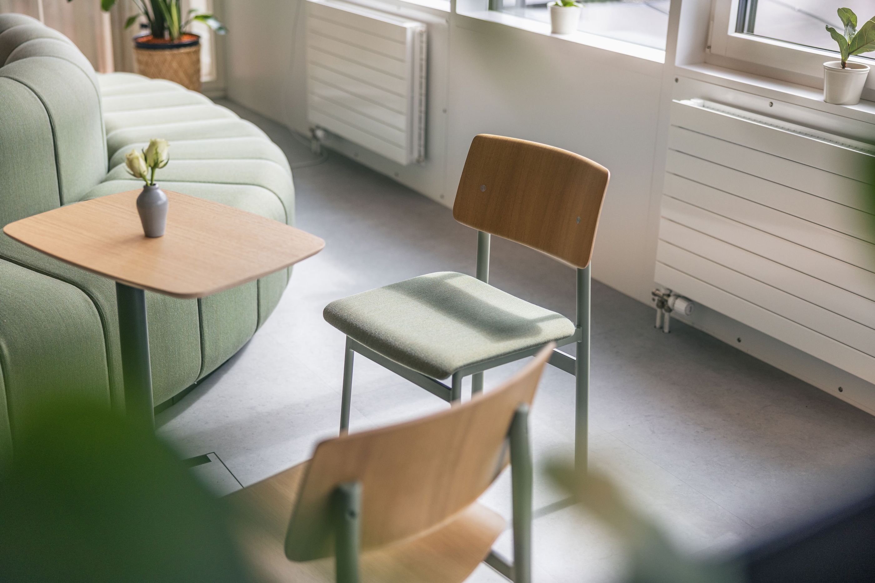 Gemütlicher Büro-Loungebereich mit grünem Sofa, Holzstühlen und kleinen Beistelltischen im Tageslicht.
