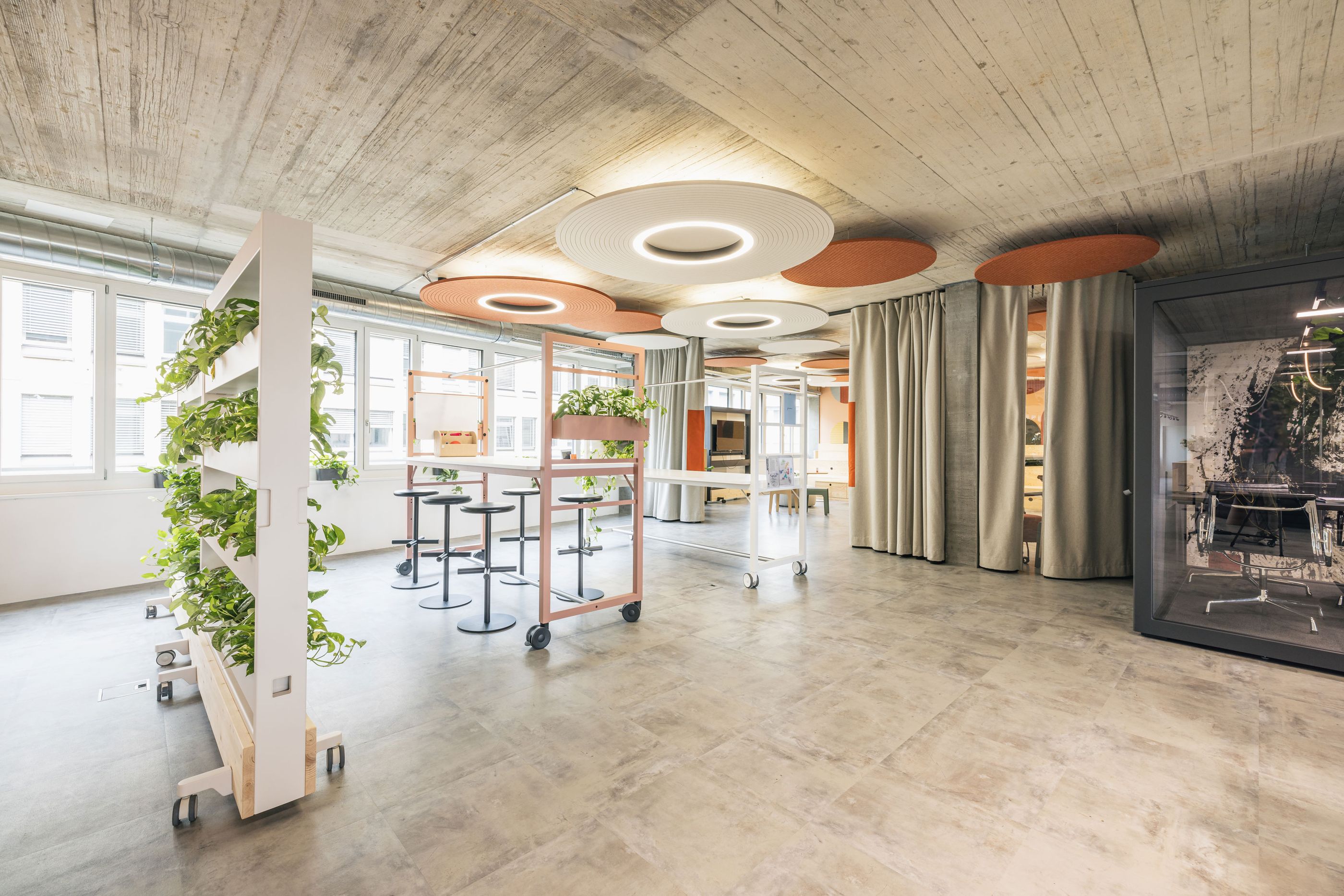 Flexibler Kreativraum mit mobilen Workshopstationen, Pflanzen und runden Akustikdeckenleuchten.
