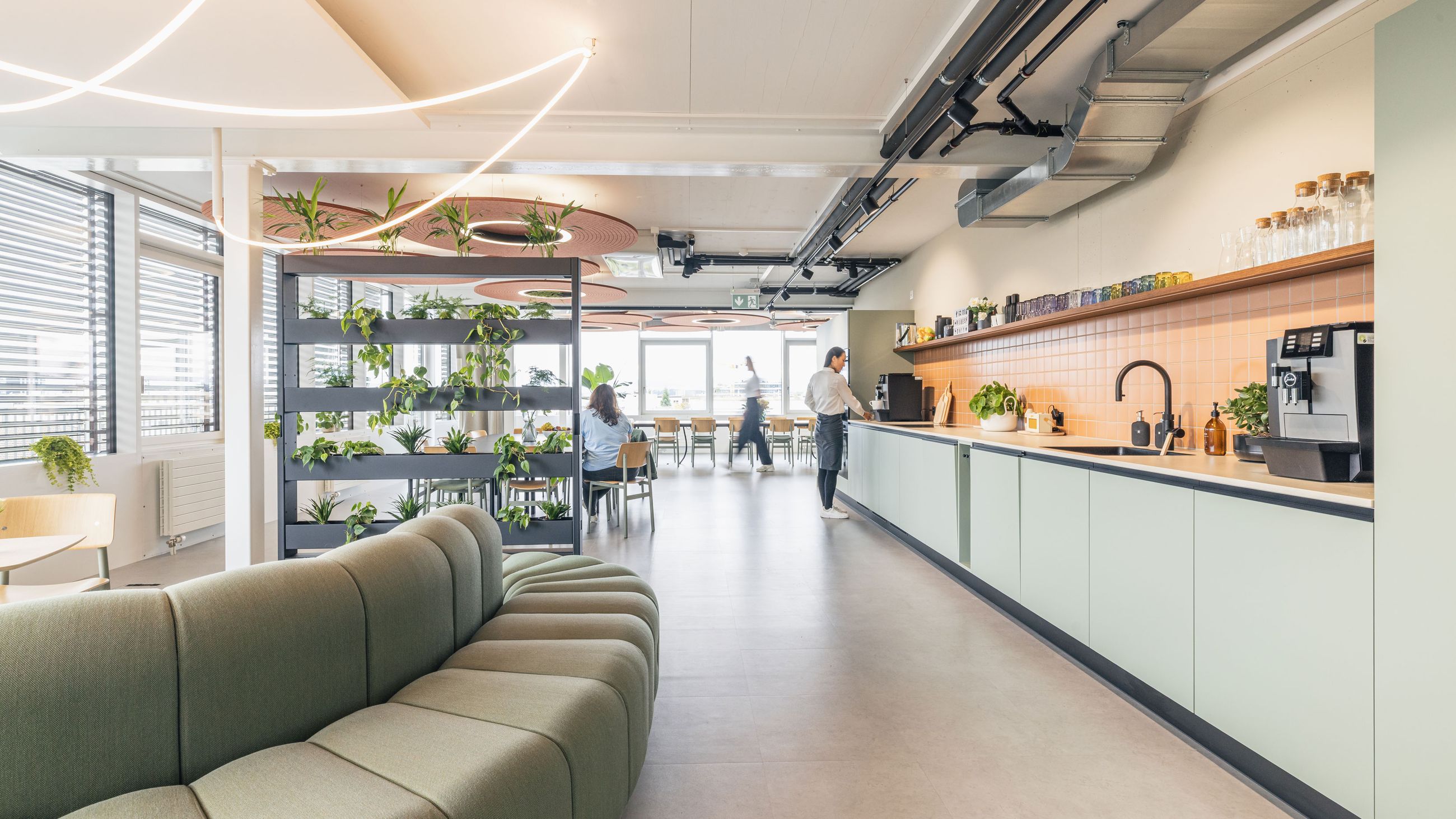 Heller Office-Community-Bereich mit Designsofa, Pflanzenregal und moderner Büroküche.