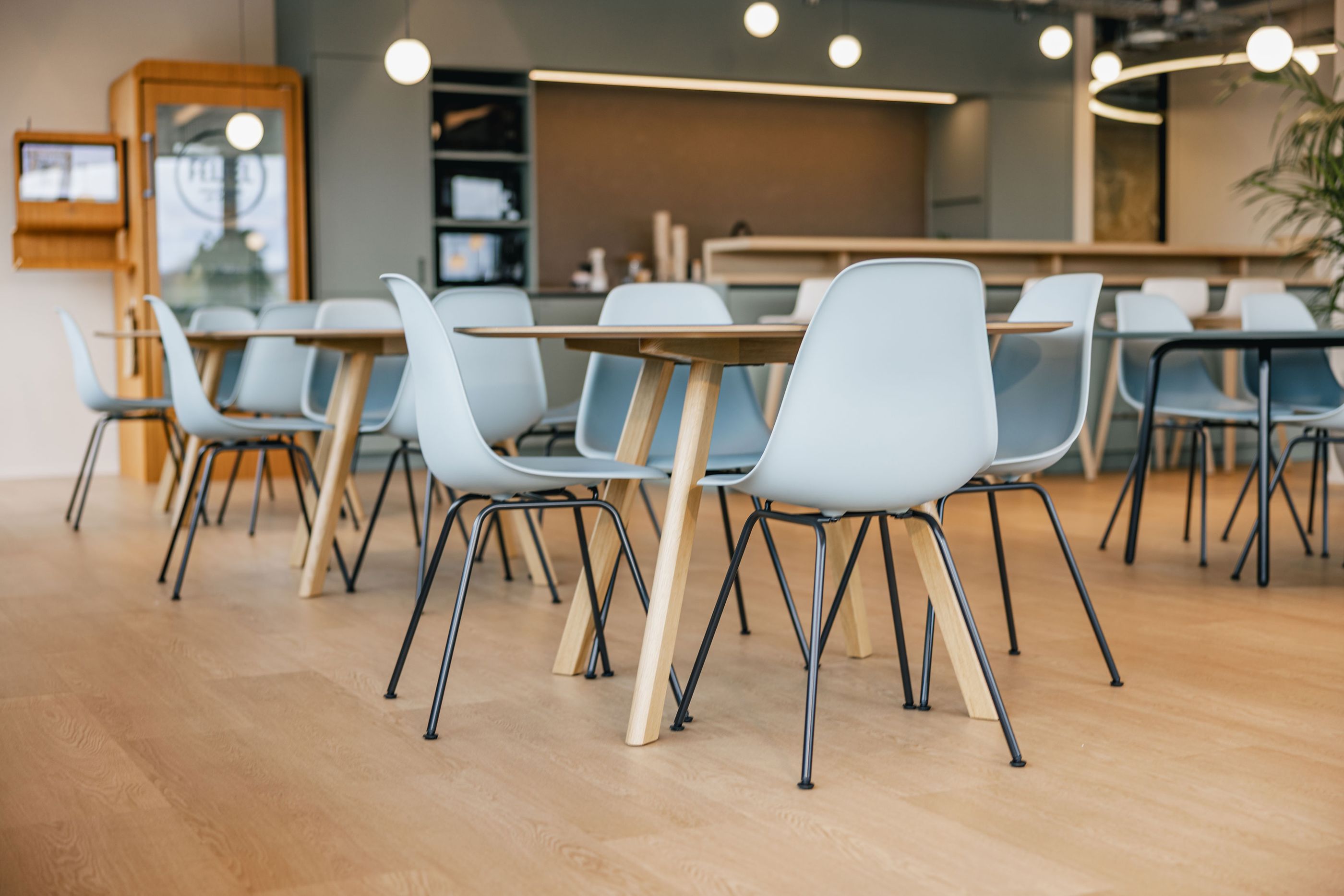 Mehrere helle Holz- und Metalltische mit pastellblauen Stühlen in einem offenen, lichtdurchfluteten Raum.
