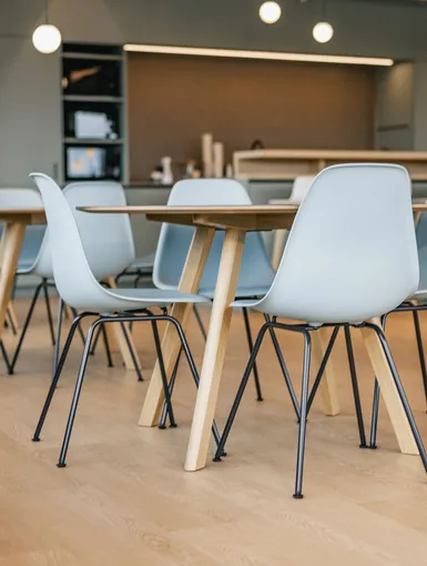 Mehrere helle Holz- und Metalltische mit pastellblauen Stühlen in einem offenen, lichtdurchfluteten Raum.