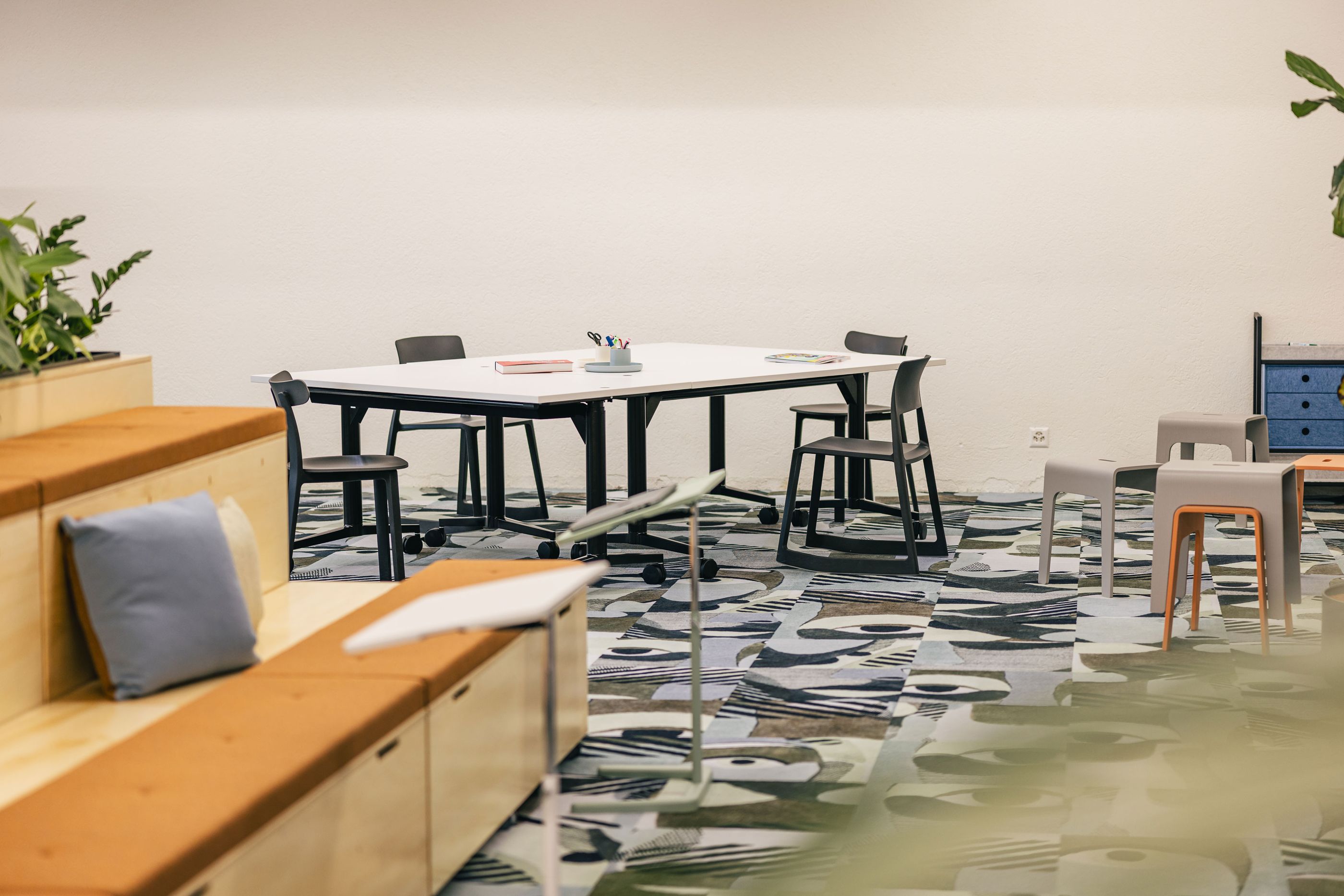 Meetingraum mit weißem Tisch, vier Stühlen und auffälligem Teppich.