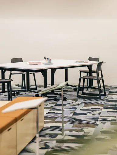 Meetingraum mit weißem Tisch, vier Stühlen und auffälligem Teppich.