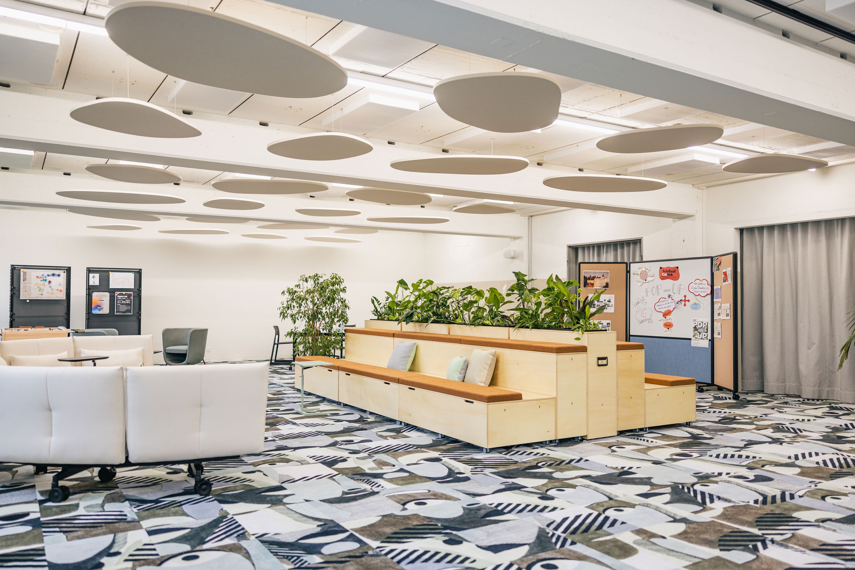 Offener Meetingbereich mit Pflanzen, Holzpodesten und großem Bildschirm.