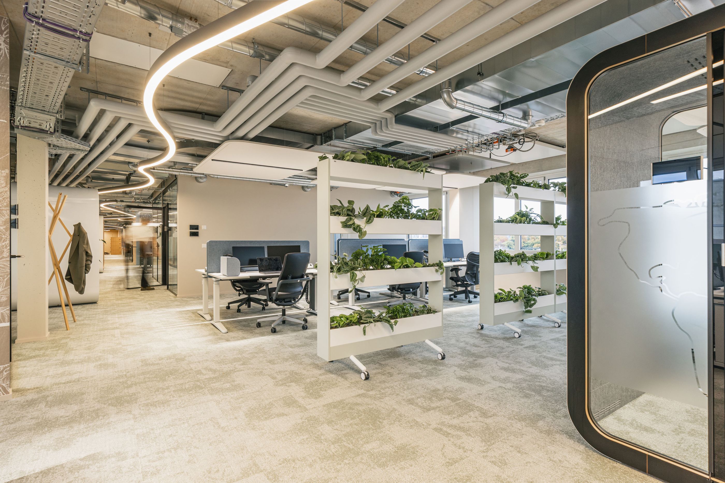 Modernes Open-Space-Büro mit begrünten Raumteilern, mehreren Arbeitsplätzen und geschwungener Deckenbeleuchtung.