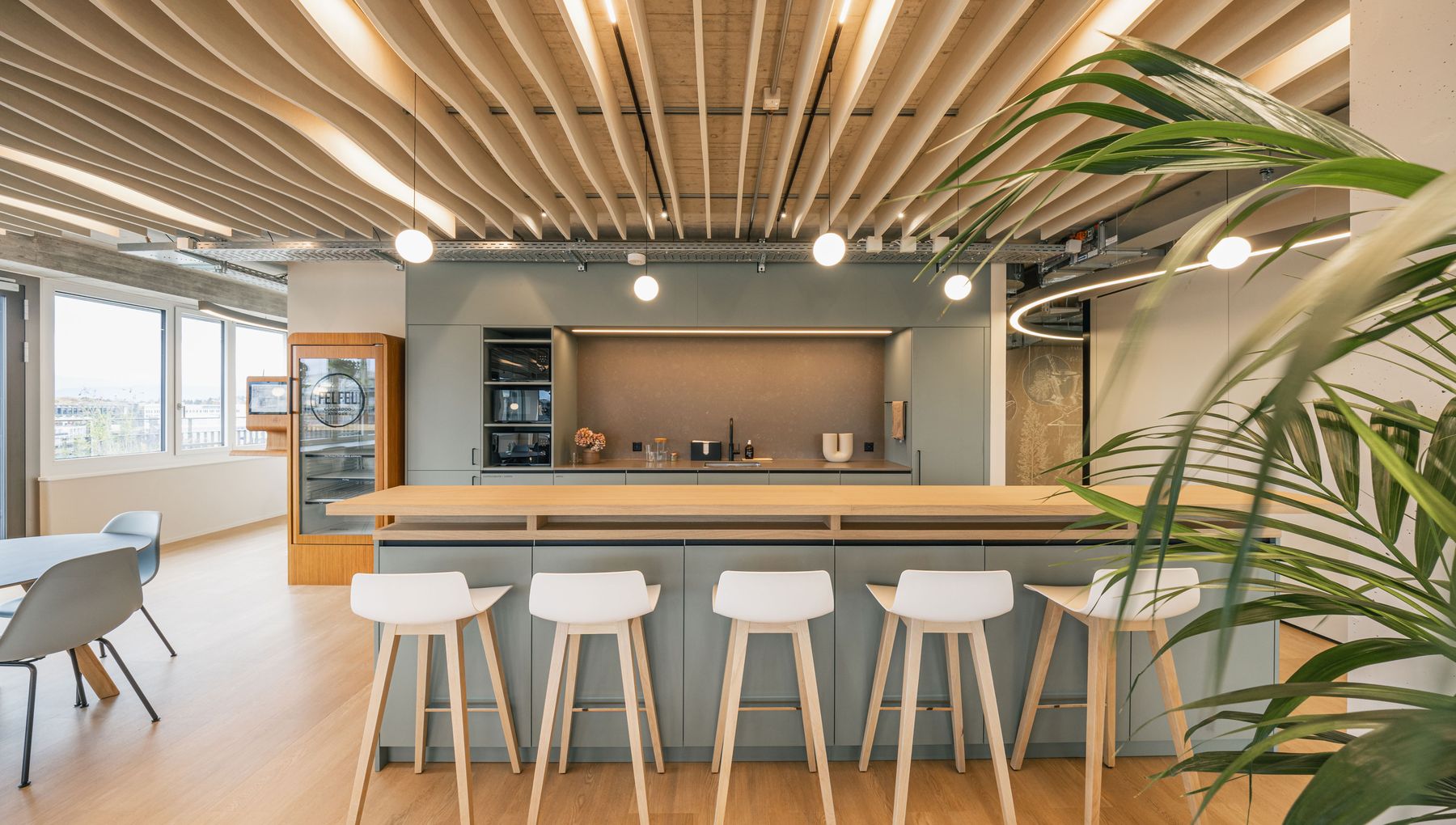 Große, moderne Büroküche mit Holztheke, mehreren weißen Barhockern und einer akzentuierten Deckenbeleuchtung.