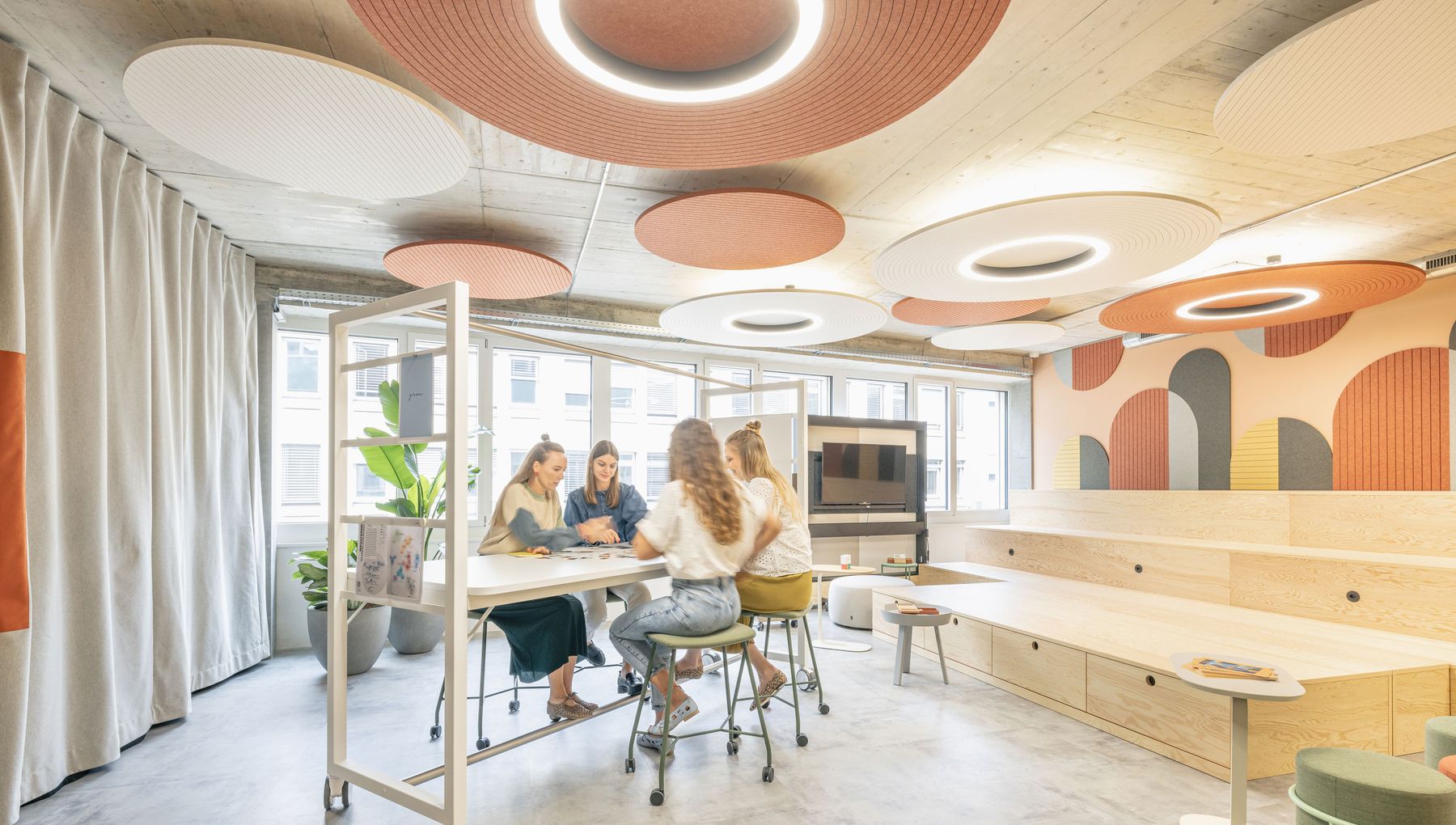 Heller Workshopraum mit runden Akustikdecken, Holzbühne und Teammeeting an einem hohen Arbeitstisch.