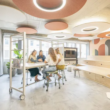 Heller Workshopraum mit runden Akustikdecken, Holzbühne und Teammeeting an einem hohen Arbeitstisch.