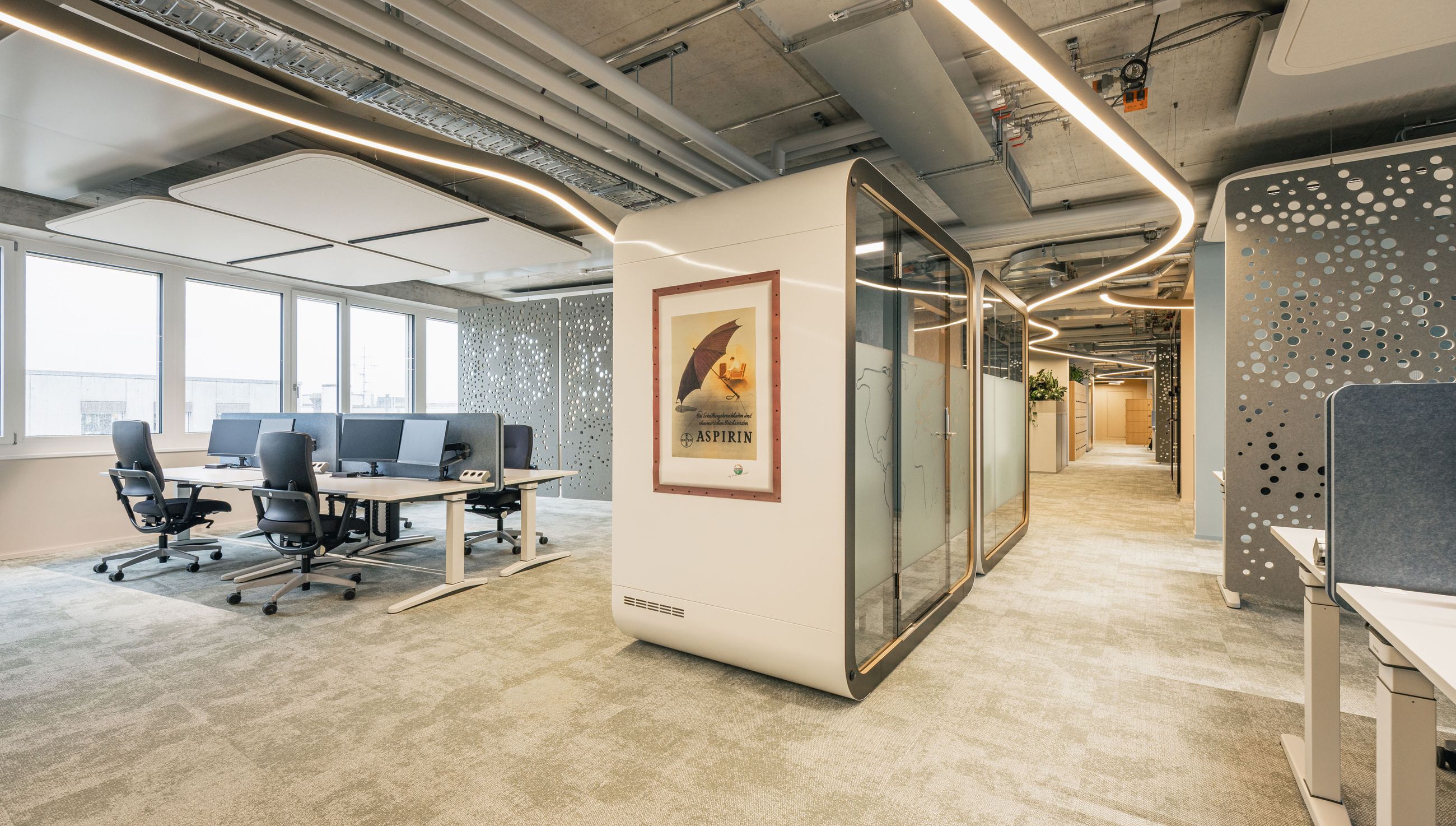 Helles Open-Space-Büro mit Schreibtischen, Telefonbox, dekorativ durchbrochenen Wandpaneelen und sichtbarer Deckeninstallation.