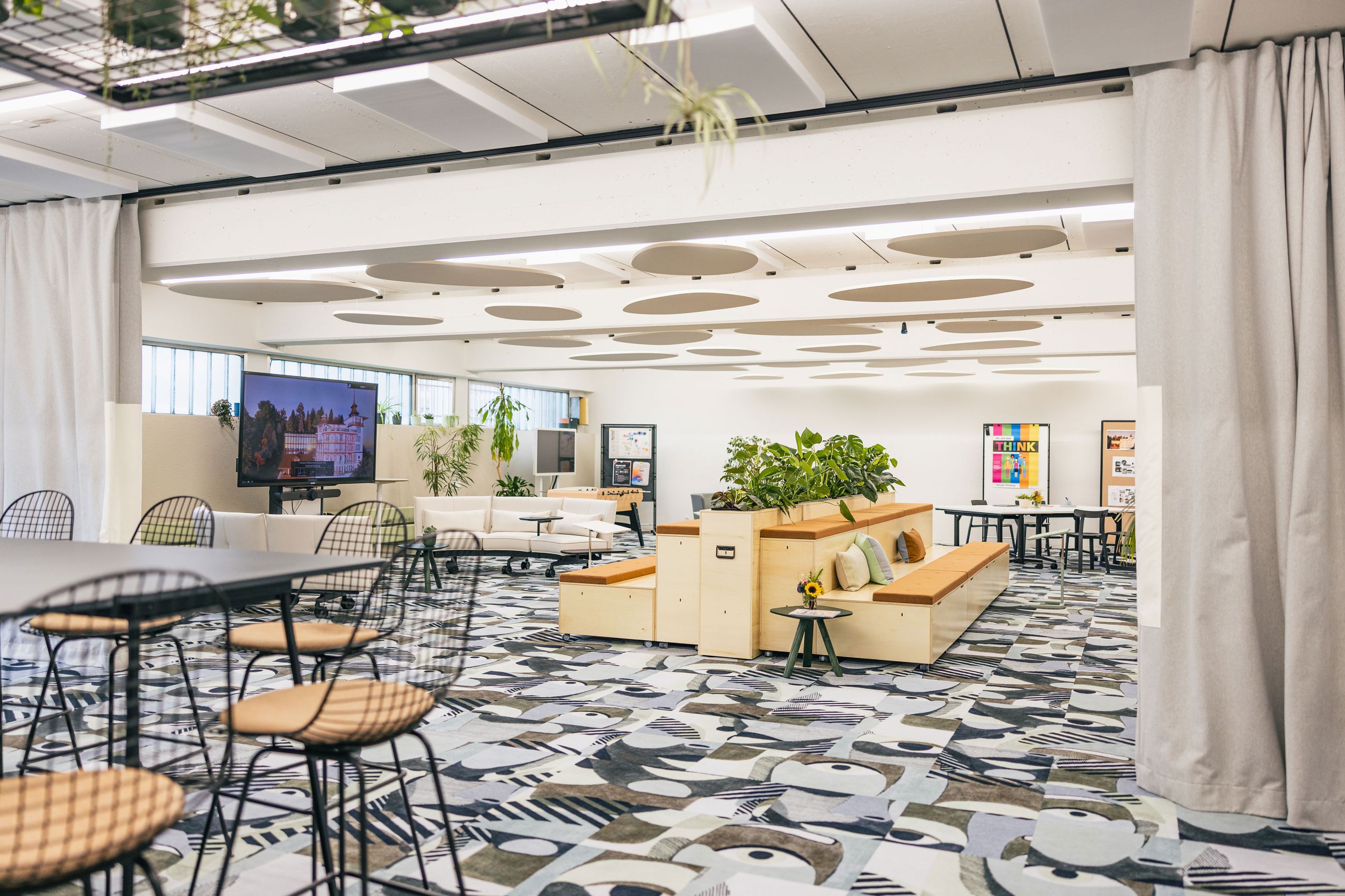 Offener Meetingbereich mit Pflanzen, Holzpodesten und großem Bildschirm.