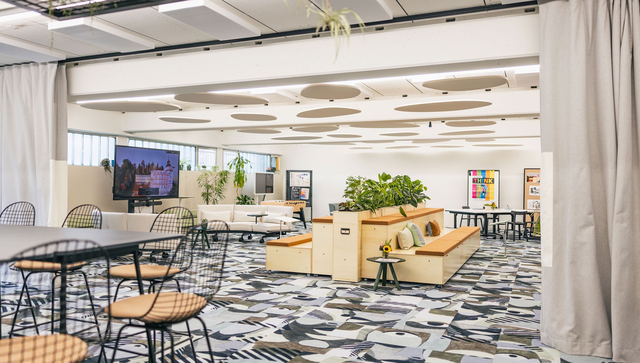 Offener Meetingbereich mit Pflanzen, Holzpodesten und großem Bildschirm.