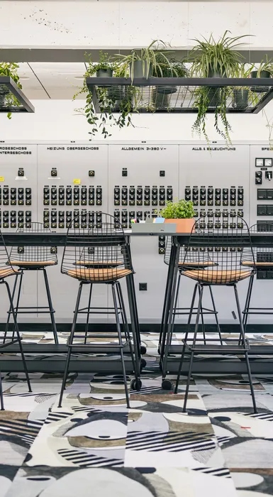 Workshopzone mit großem Tisch, schwarzen Stühlen und farbigem Teppich.