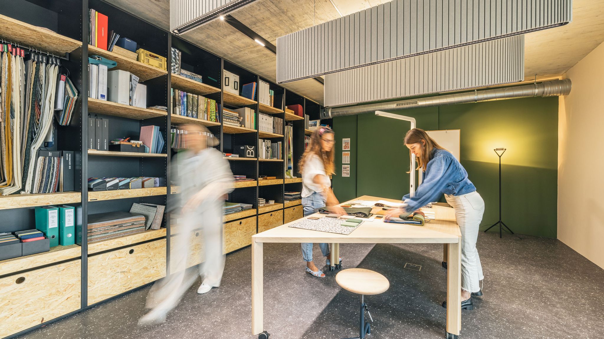 Kreativer Materialraum mit großen Regalen, Stoffmustern und Teamwork an einem zentralen Arbeitstisch.