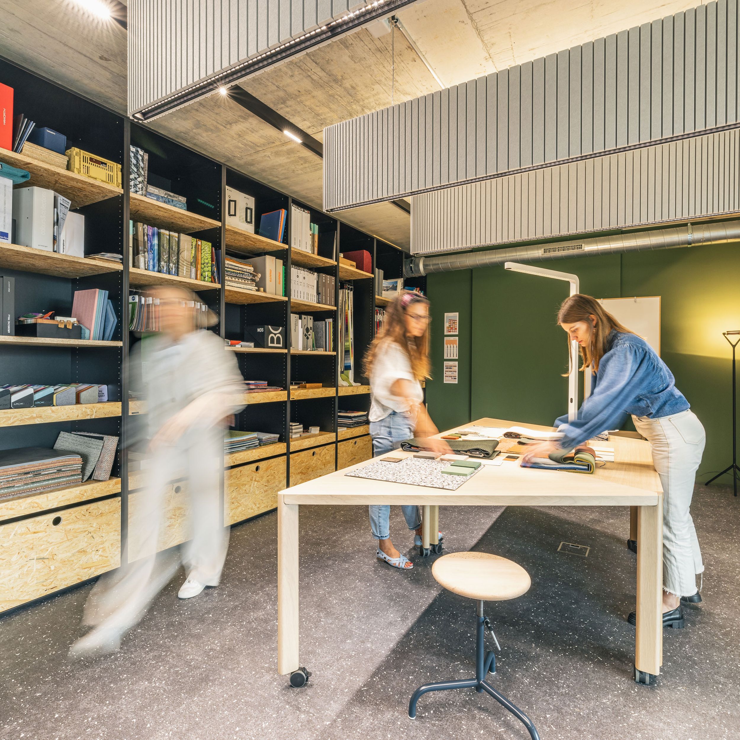 Kreativer Materialraum mit großen Regalen, Stoffmustern und Teamwork an einem zentralen Arbeitstisch.