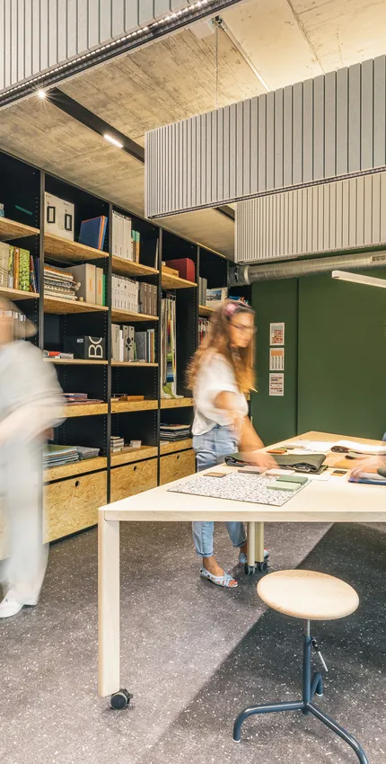 Kreativer Materialraum mit großen Regalen, Stoffmustern und Teamwork an einem zentralen Arbeitstisch.