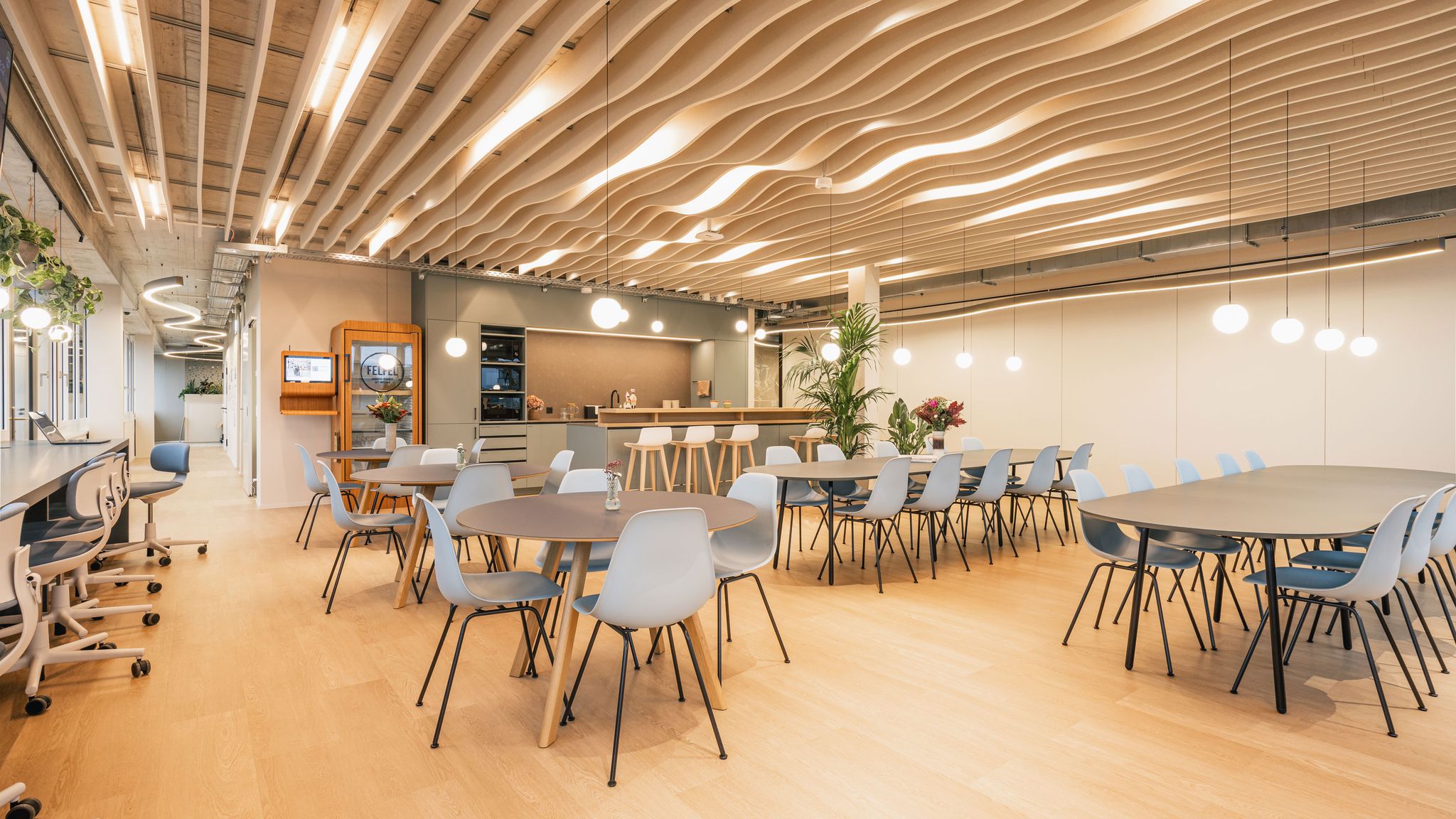 Großer Aufenthaltsbereich mit Tischen, hellblauen Stühlen, offener Küche und wellenförmiger Decke.