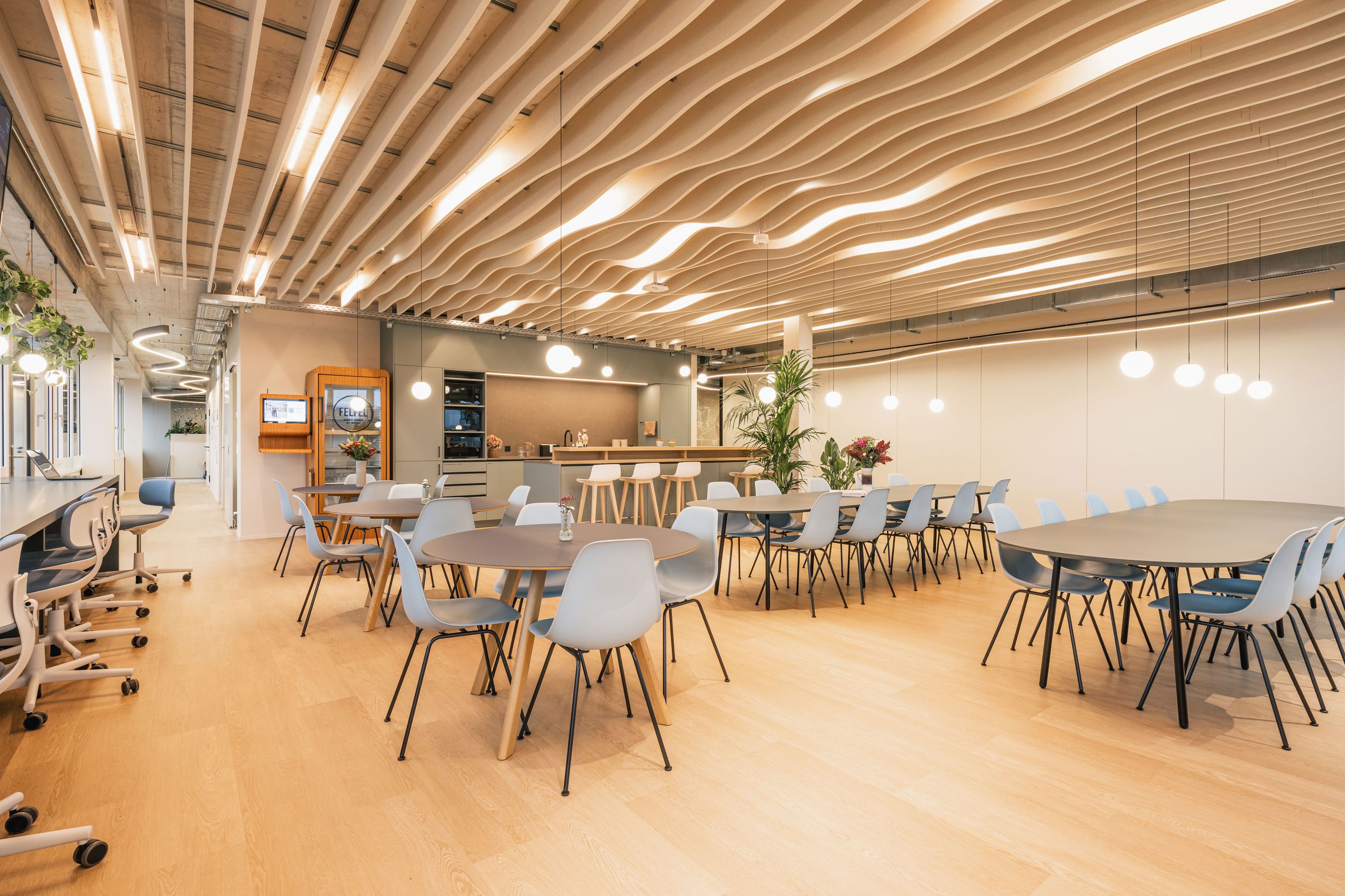 Großer Aufenthaltsbereich mit Tischen, hellblauen Stühlen, offener Küche und wellenförmiger Decke.