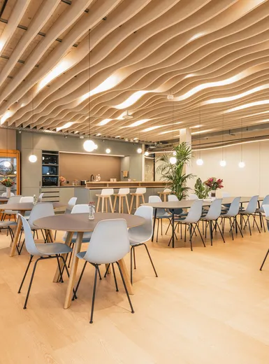 Großer Aufenthaltsbereich mit Tischen, hellblauen Stühlen, offener Küche und wellenförmiger Decke.