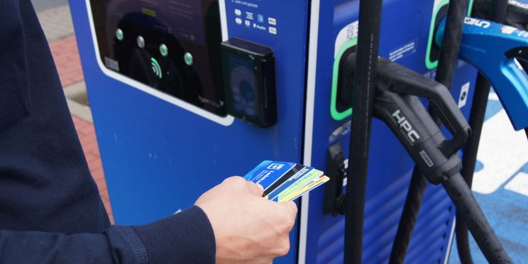 Person steht vor einer blauen Ladestation und hält mehrere Ladekarten in der Hand.