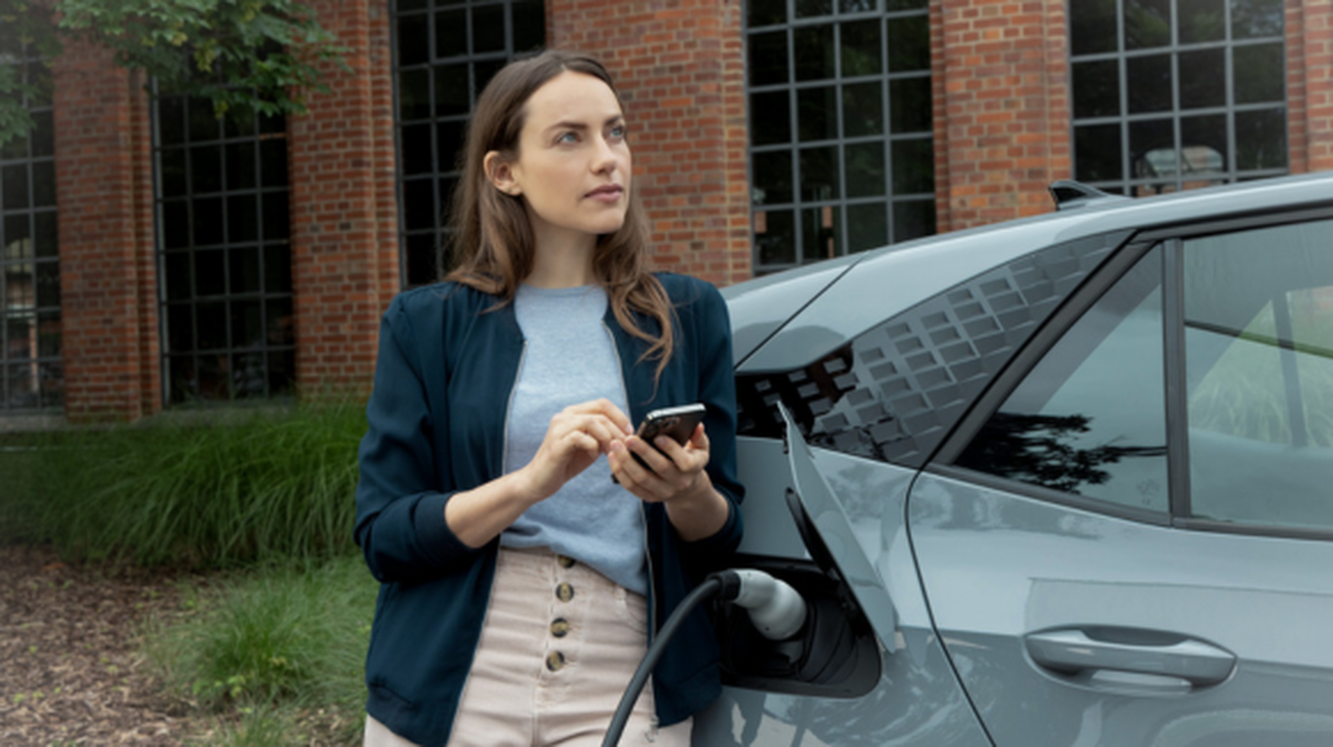 Frau hält Smartphone in der Hand, während e-Auto lädt.