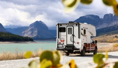 Camper van driving along a lakeside road with mountains in the background, framed by leafy branches.