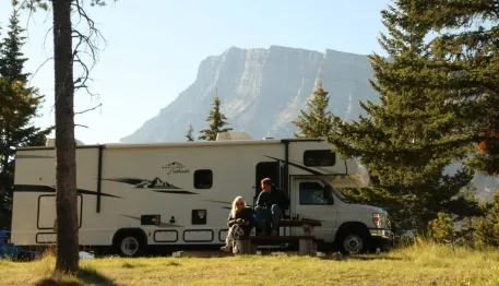 An RV parked in a scenic campsite with two people sitting at a picnic table. Pine trees and a mountain provide a stunning backdrop.