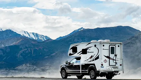 Pickup truck with camper parked on a road, set against a backdrop of majestic mountains and a cloudy sky.