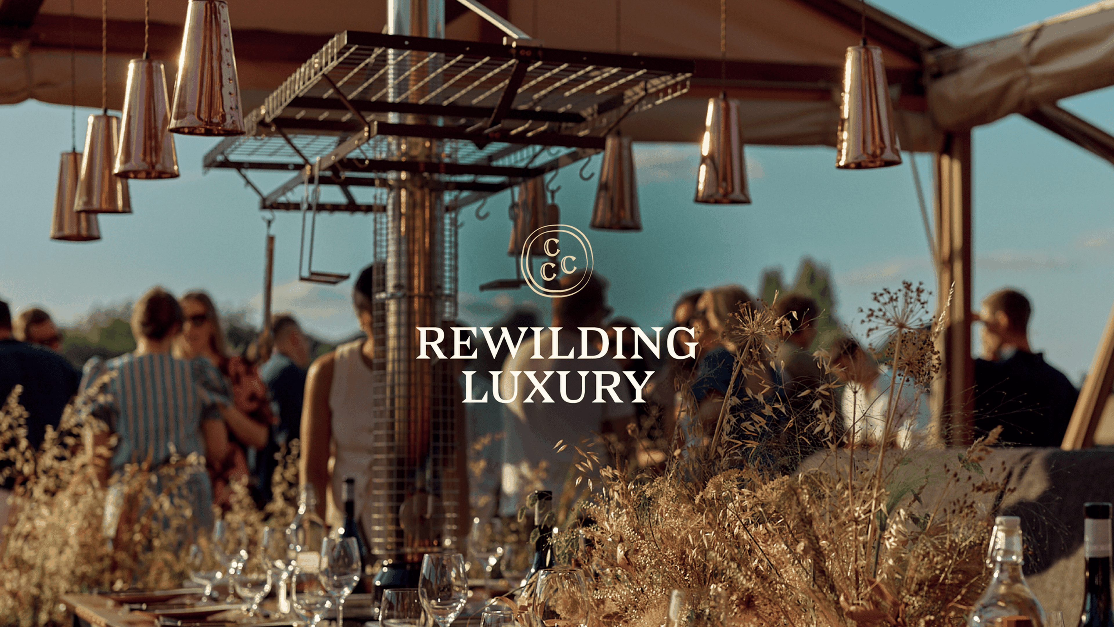 Outdoor gathering under a tent with copper lights, rustic decor, glasses, and "Rewilding Luxury" text overlay in the center.