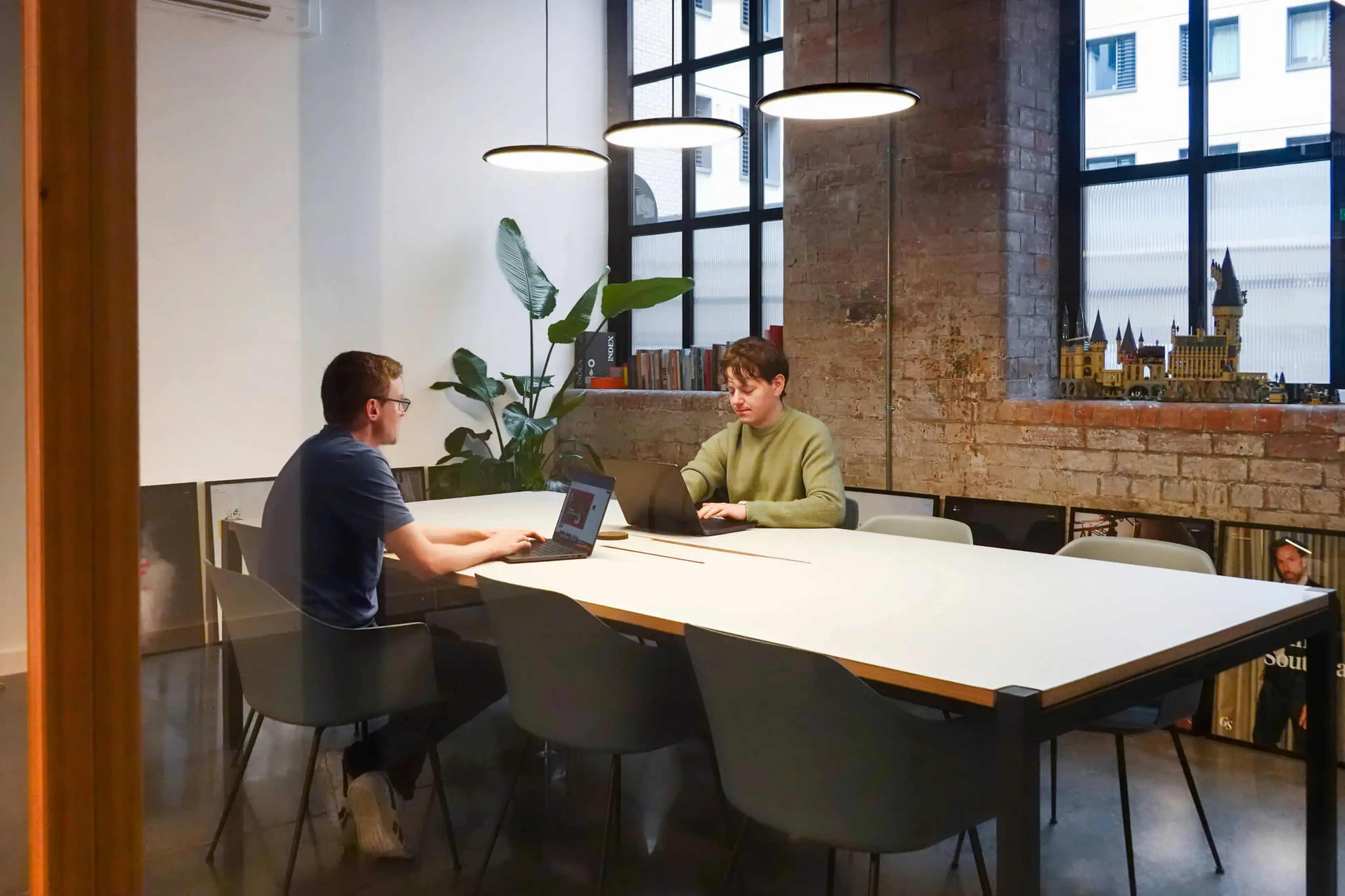 Two people are working on laptops at a rectangular table in a modern office with large windows and a brick wall.