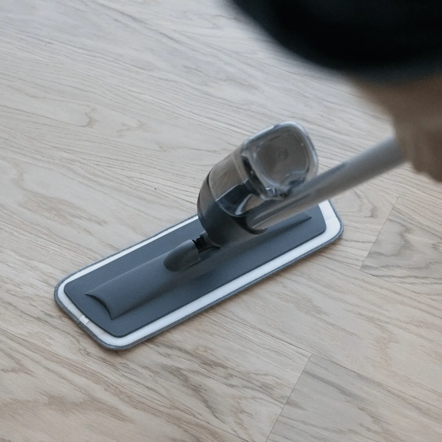 A close-up of a person using a spray mop on a light wood floor, with a focus on the mop's head and handle.