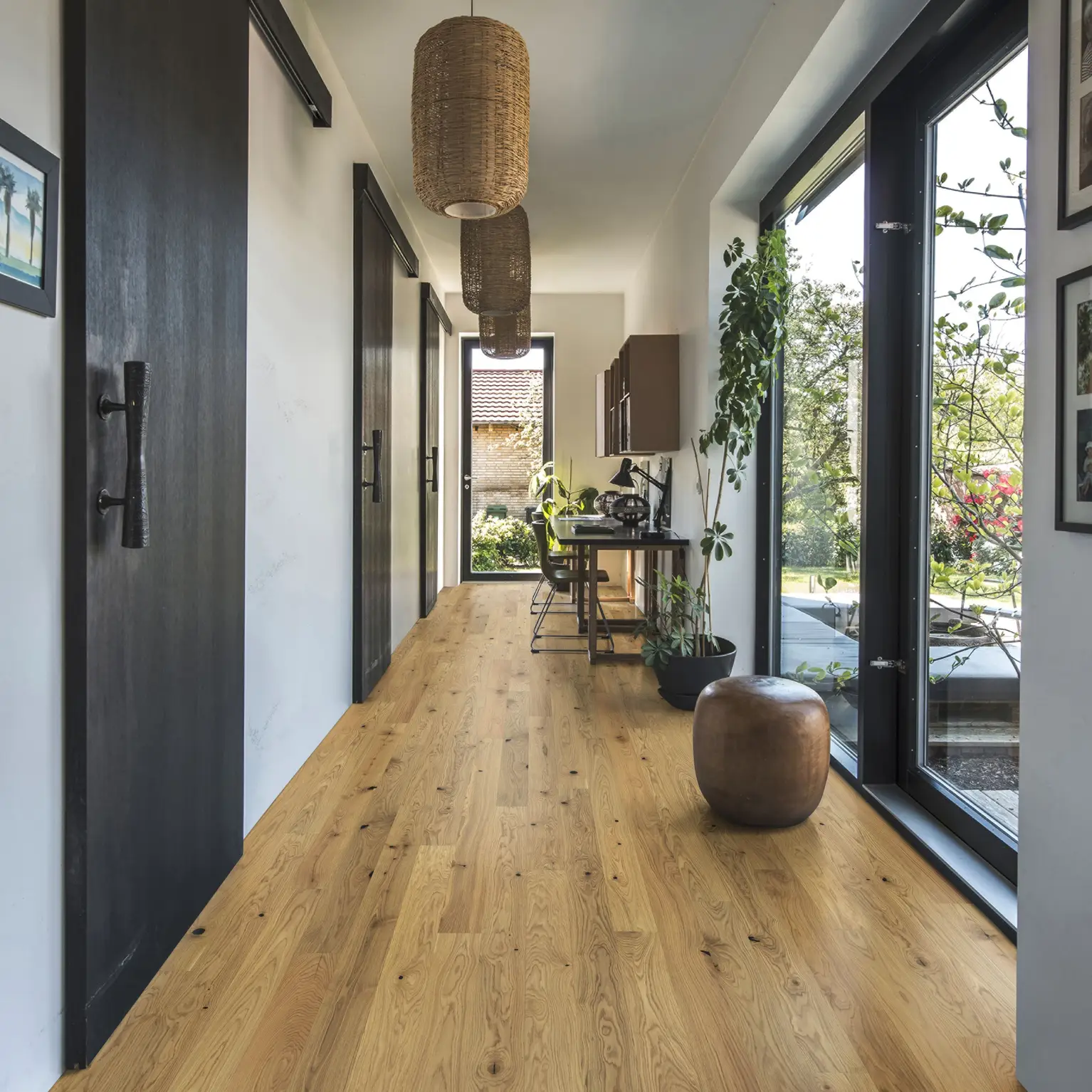 A hallway with warm oak flooring, complemented by abundant greenery, woven pendant lights, and large windows letting in natural light.