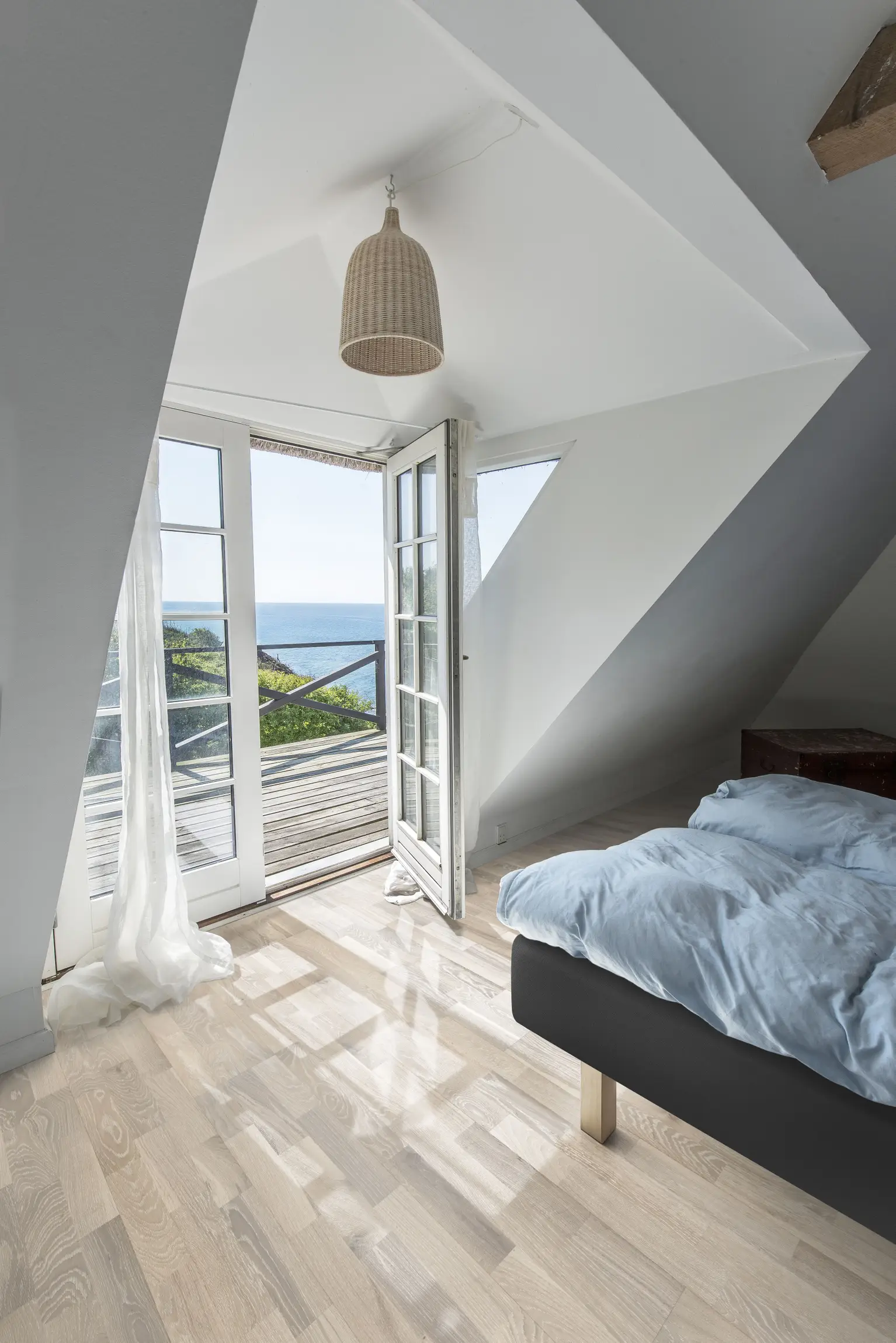 Bright bedroom with wooden floor, blue bedding, and open glass doors leading to a balcony with ocean view. Hanging wicker lamp adds a cozy touch.