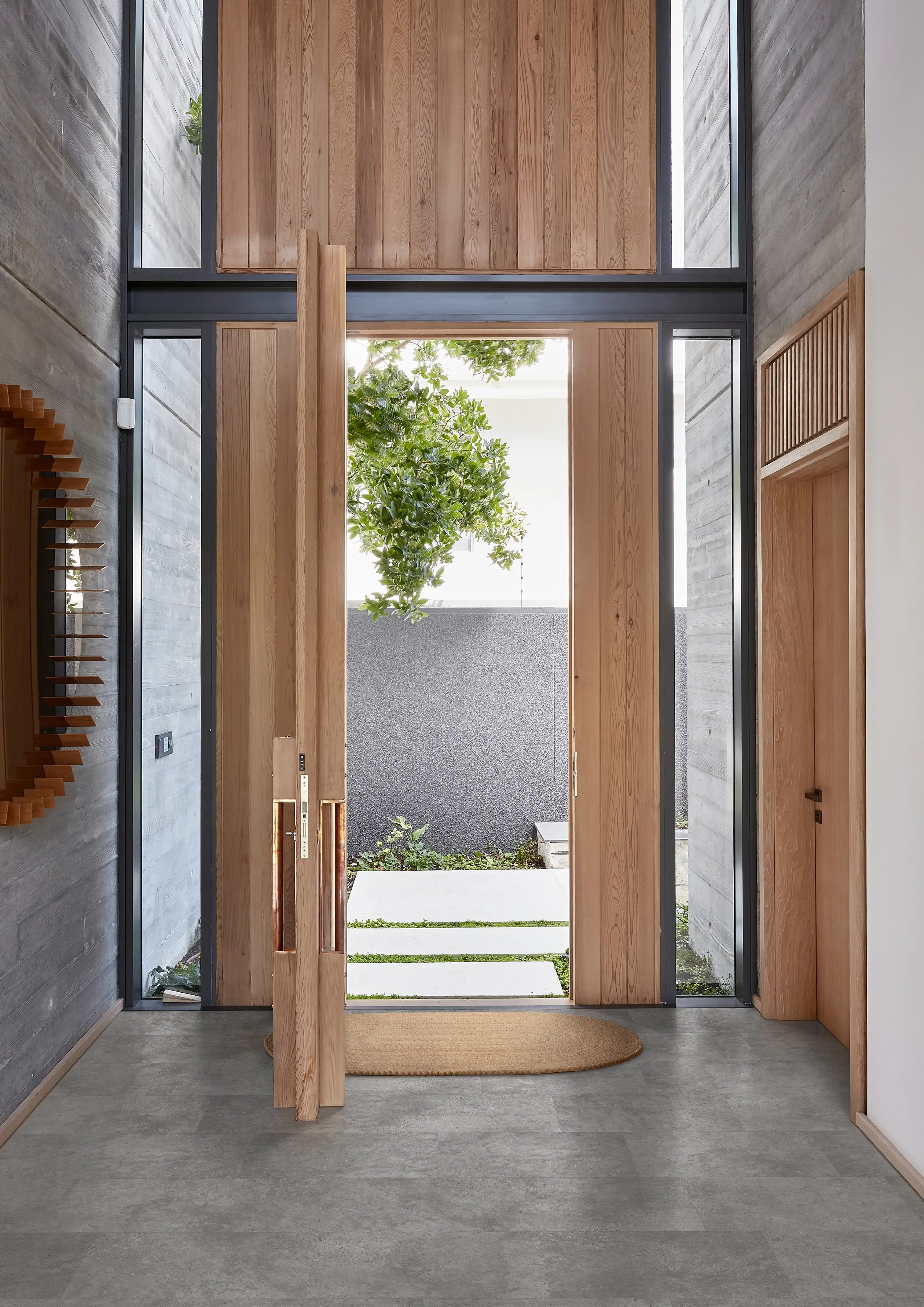 Modern entryway with tall wooden doors, concrete walls, and a view of a small garden outside. A round mirror hangs on the left wall.