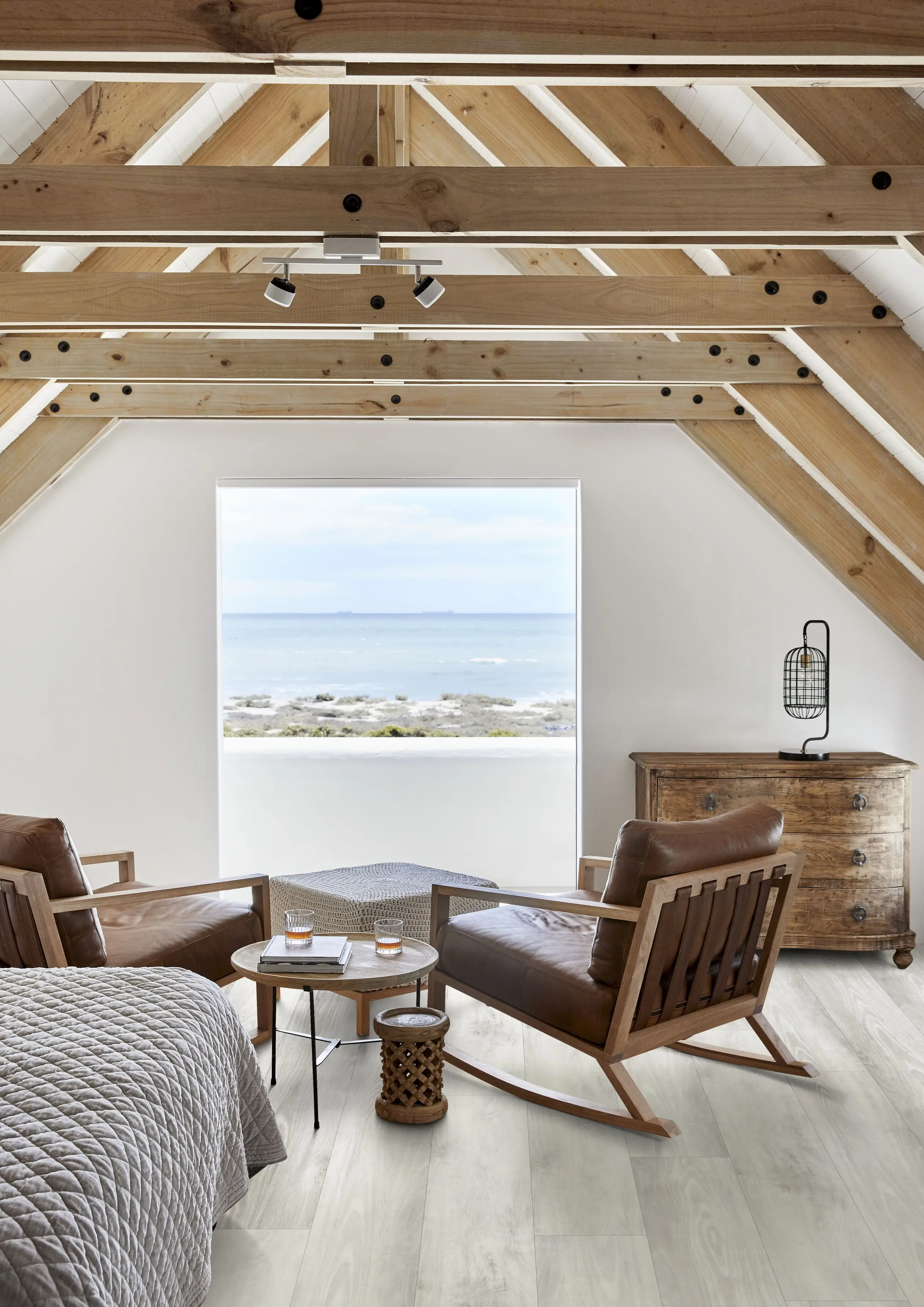 Cozy attic room with wooden beams, leather chairs, and a sea view through a large window. A bed and wooden chest complete the decor.
