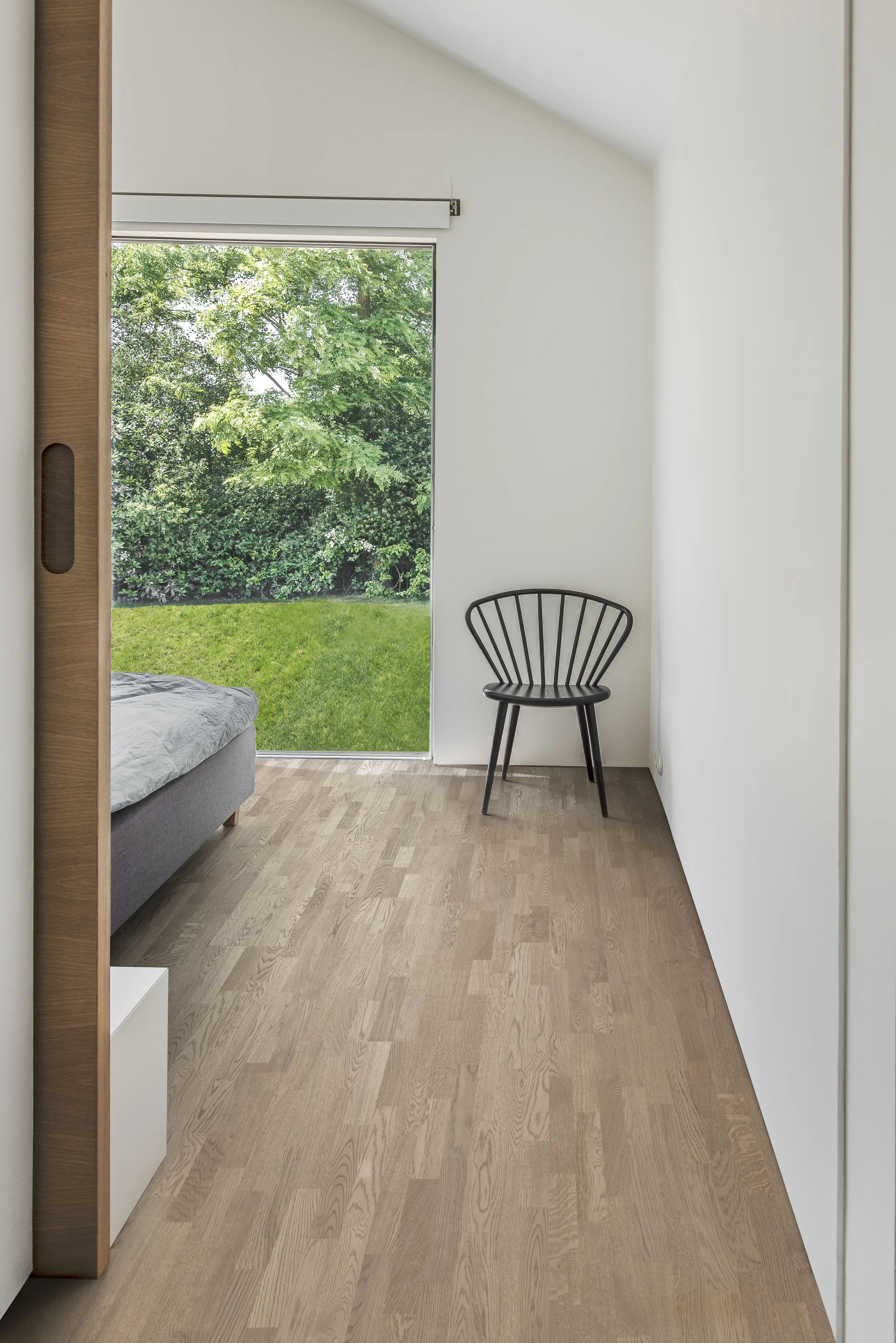 Minimalist bedroom with wooden floor, a gray bed, and a black chair. Large window shows green garden outside. White walls and sliding door.