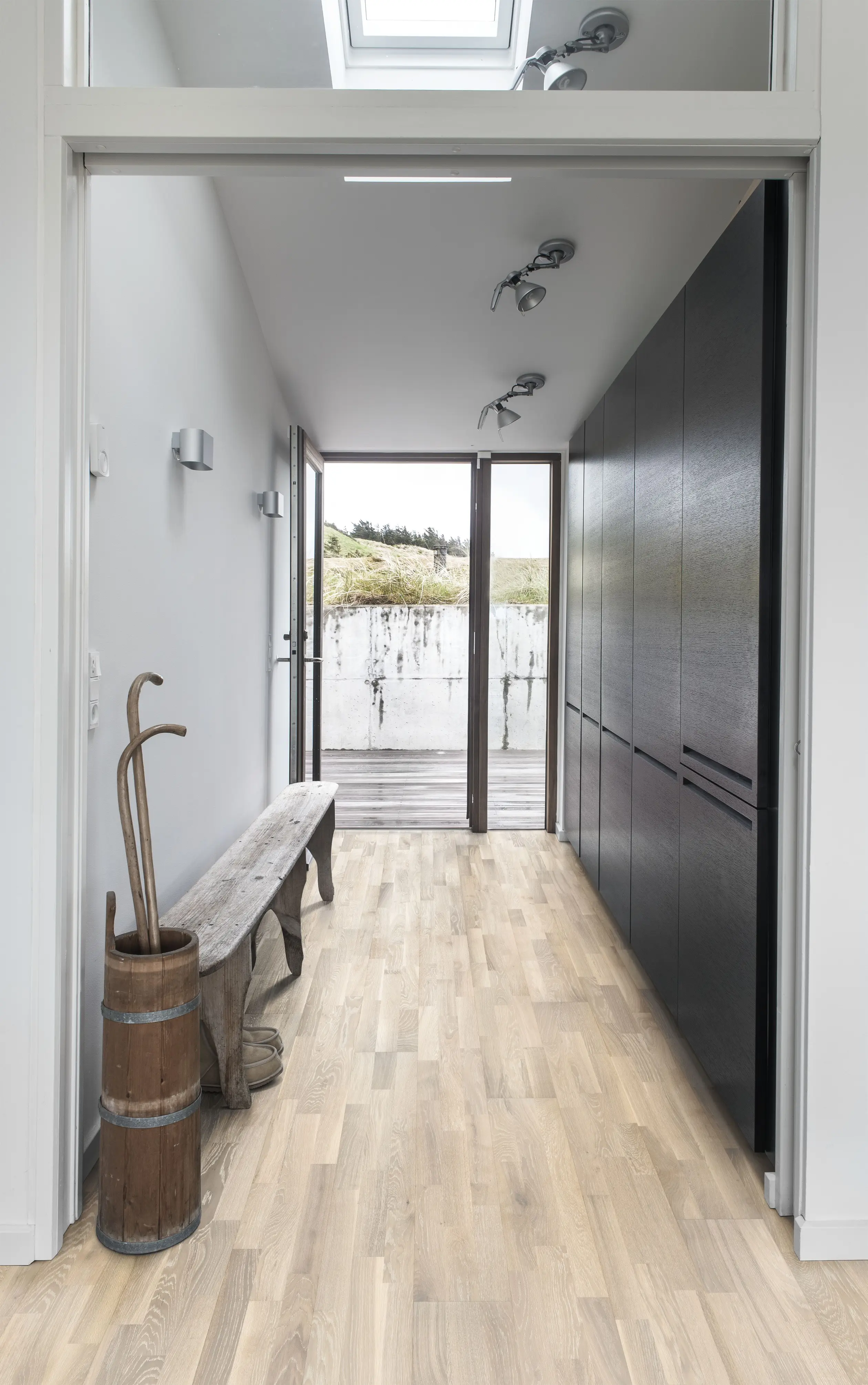 Minimalist hallway with light wood flooring, black cabinets, a rustic wooden bench, and a barrel with walking sticks. Glass door opens to a patio.