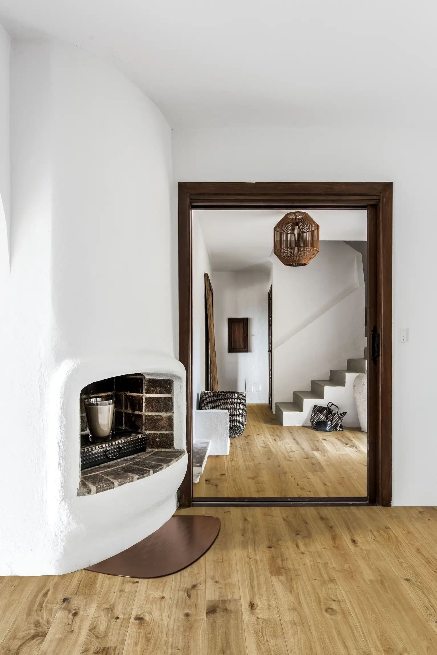 Oak vedbo. Minimalist interior with a white curved fireplace, wooden floor, and open doorway leading to a staircase. A pendant light hangs above.