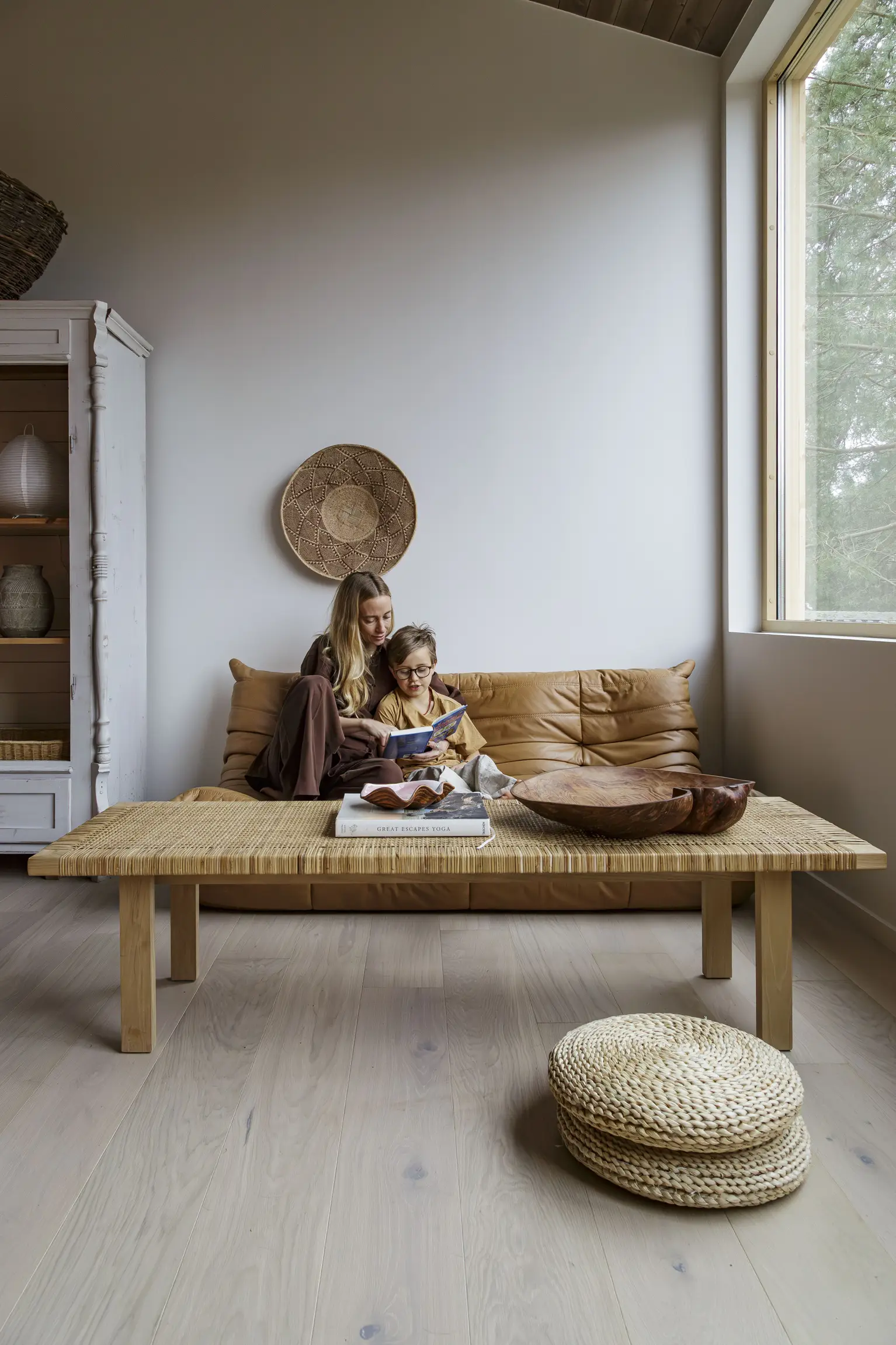 Oak loft white plank. Two people sitting on a tan couch in a minimalist living room with a wooden table, woven decor, and a large window showing trees outside.