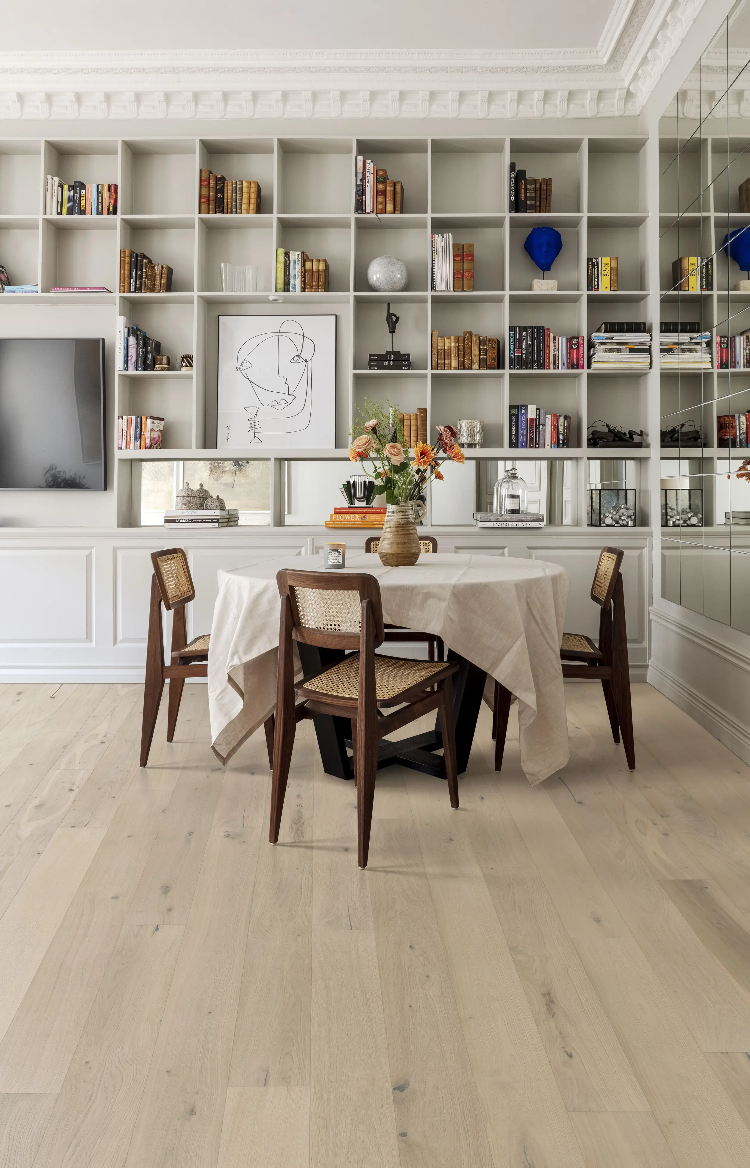 Modern dining room with a white table, wooden chairs, and a bookshelf filled with books and decor. Light wood flooring and a vase with flowers.