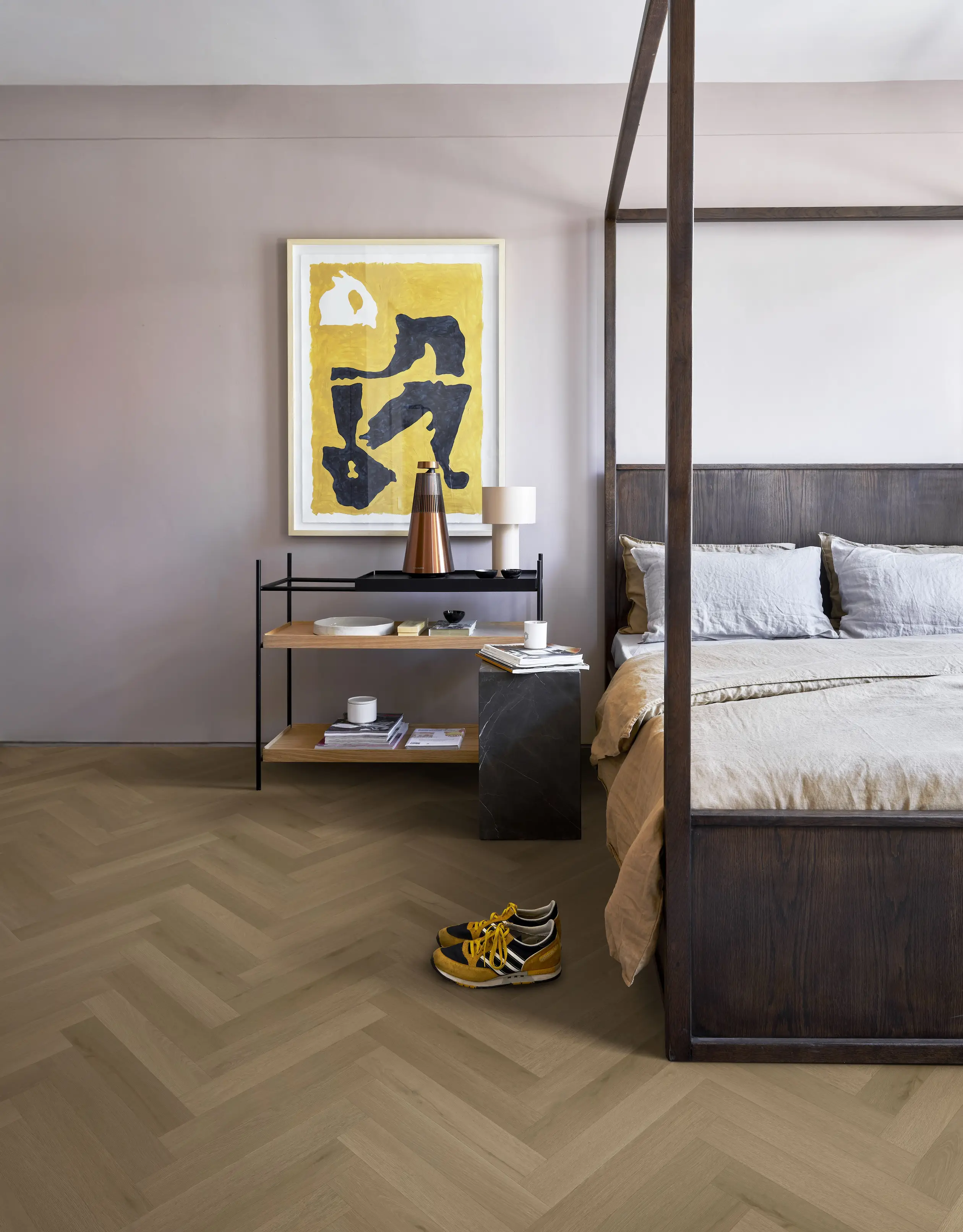 Minimalist bedroom with a wooden canopy bed, abstract art, and a side table. Yellow shoes are on the chevron-patterned floor.