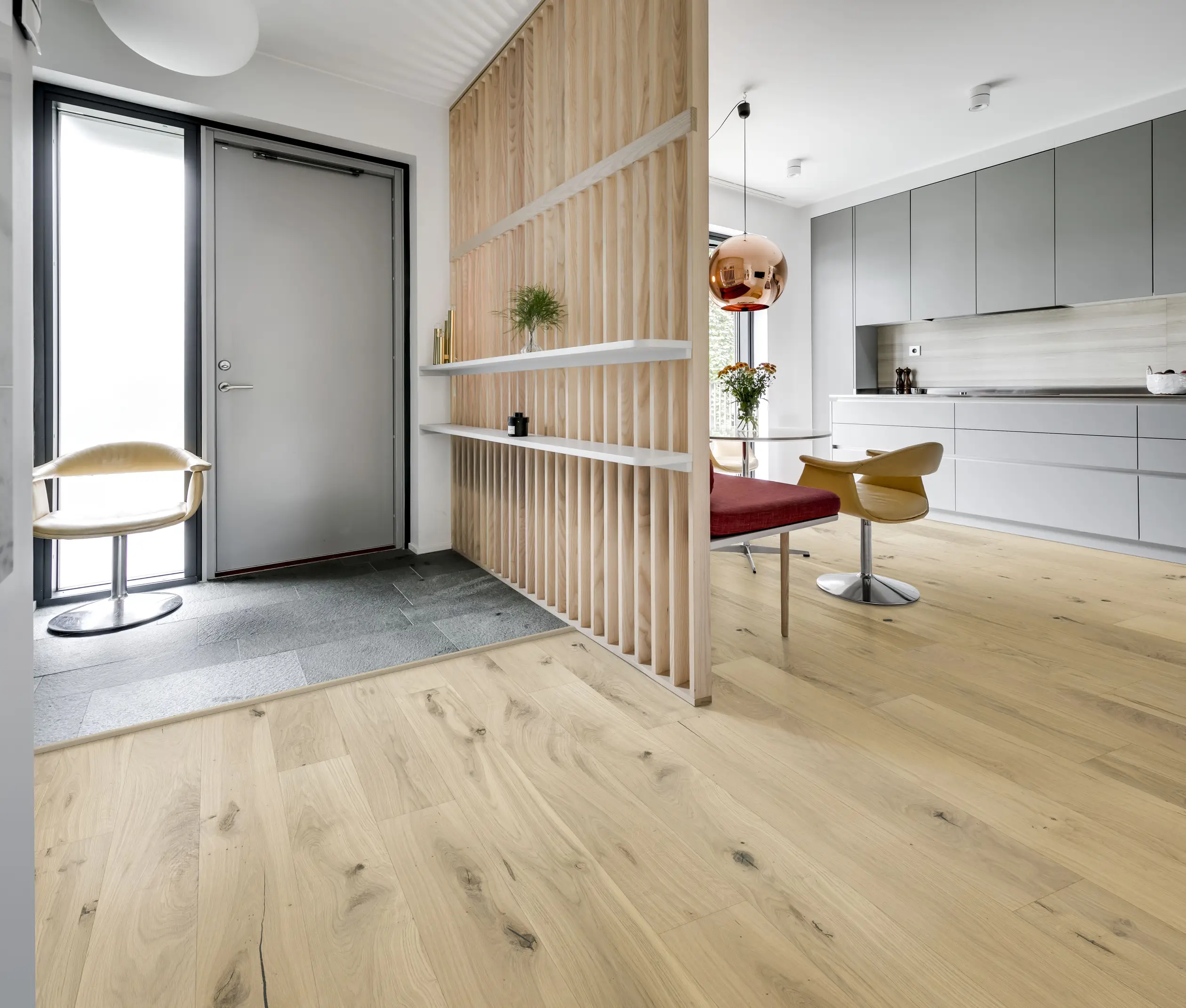 Modern open-plan interior with light wood flooring, a partition wall, gray cabinets, a red bench, and a hanging copper light fixture.