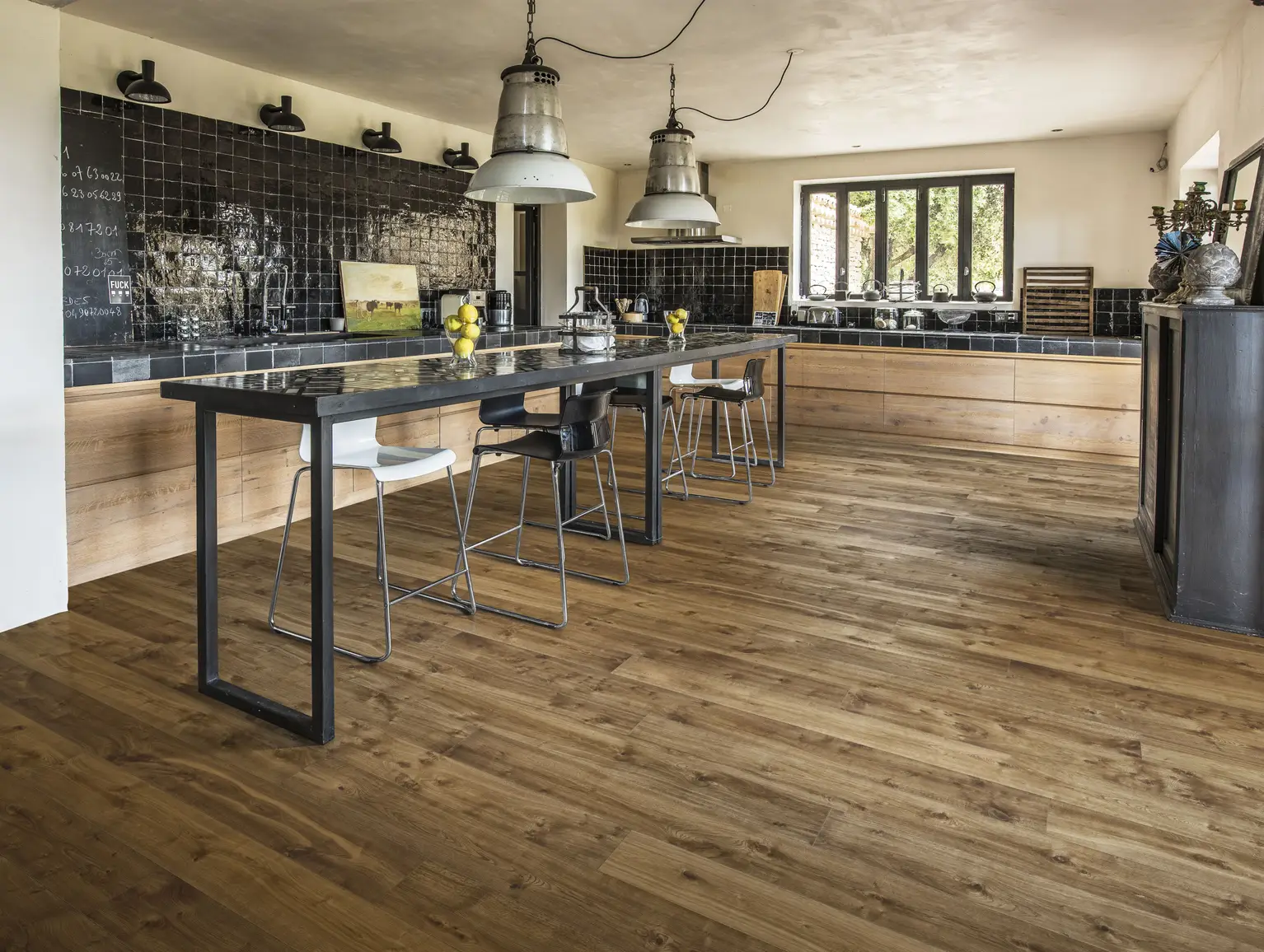 Spacious kitchen with wooden floors, a long island with bar stools, pendant lights, and a large window overlooking greenery.