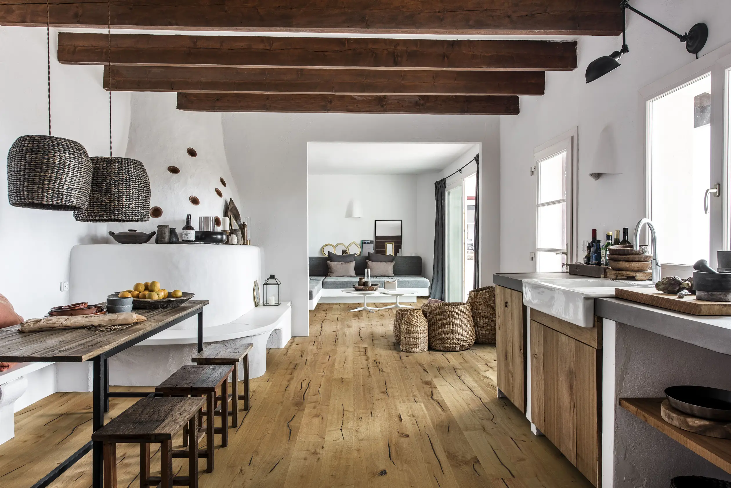 Rustic kitchen with wooden beams, a long dining table, woven pendant lights, and a cozy living area with natural light and neutral tones.